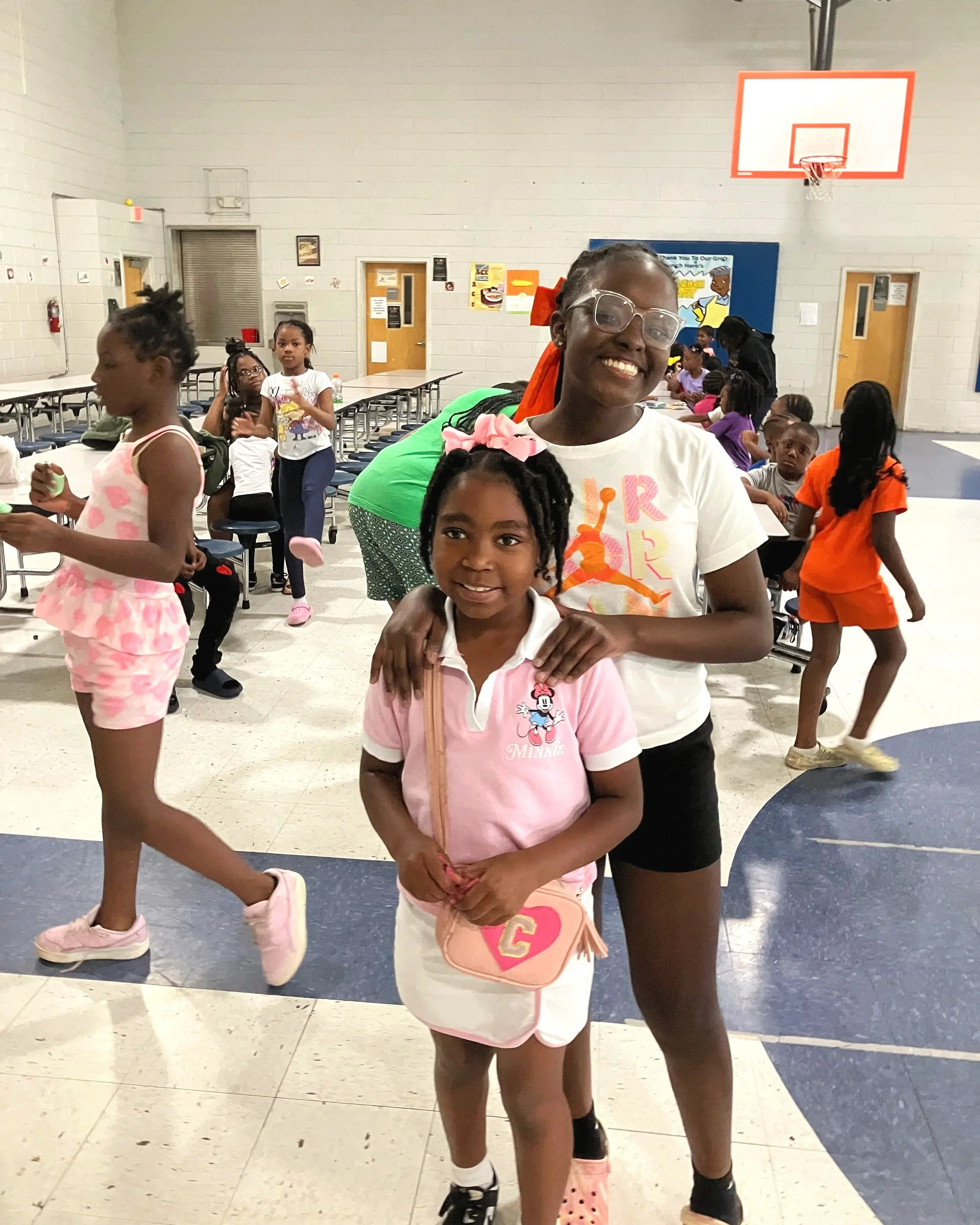 Two young girls are posing together in the foreground of what appears to be a school gymnasium or cafeteria. Many other children are visible in the background, engaged in various activities.