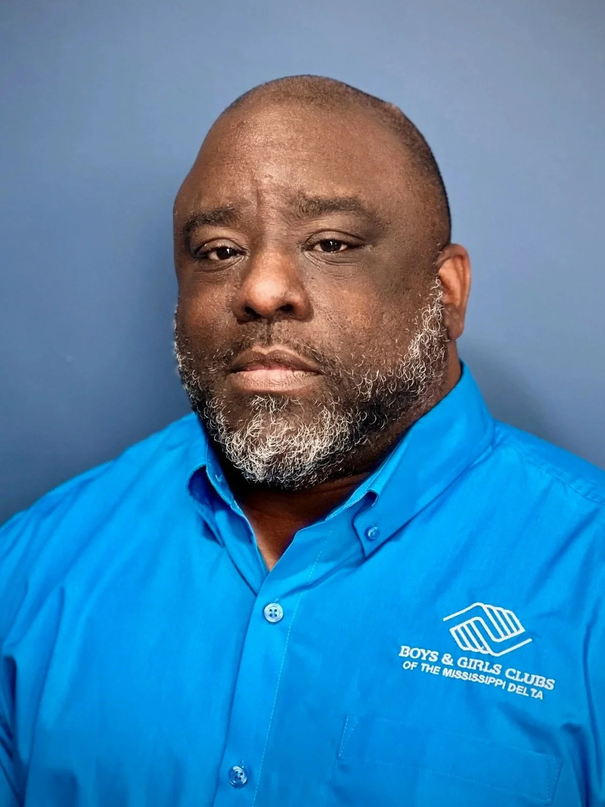A man with a short beard and a bald head wearing a blue shirt with the logo of Boys & Girls Clubs of the Mississippi Delta on it, standing against a blue background.