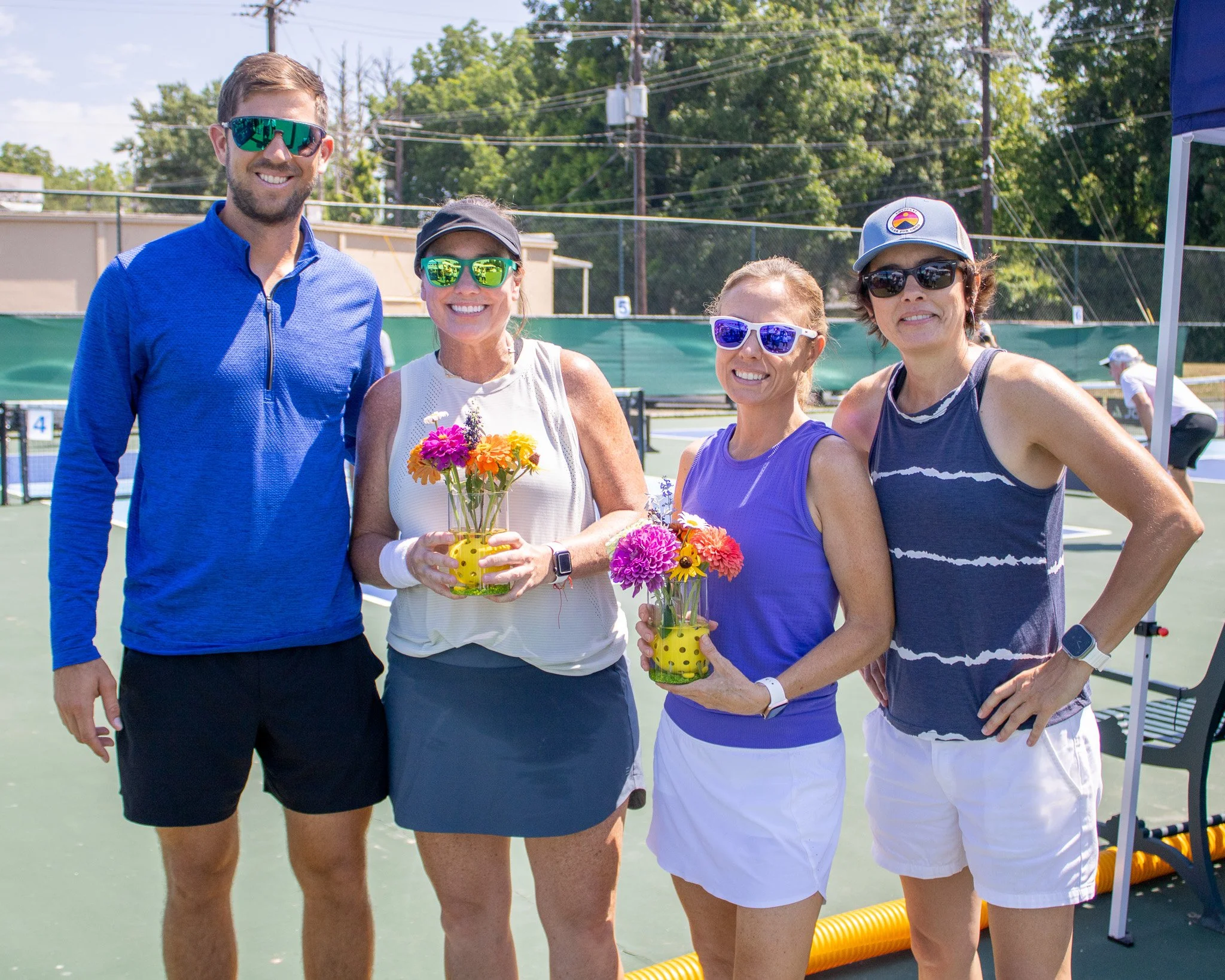Four people standing on a tennis court outdoors, wearing sporty attire and sunglasses, holding colorful flower arrangements, smiling for the photo.