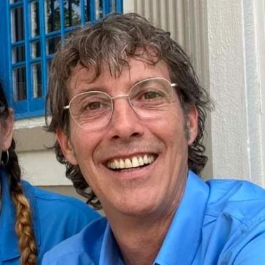 Smiling man with glasses and wavy hair wearing a blue shirt, posed outdoors near a building with a blue window and white wall.