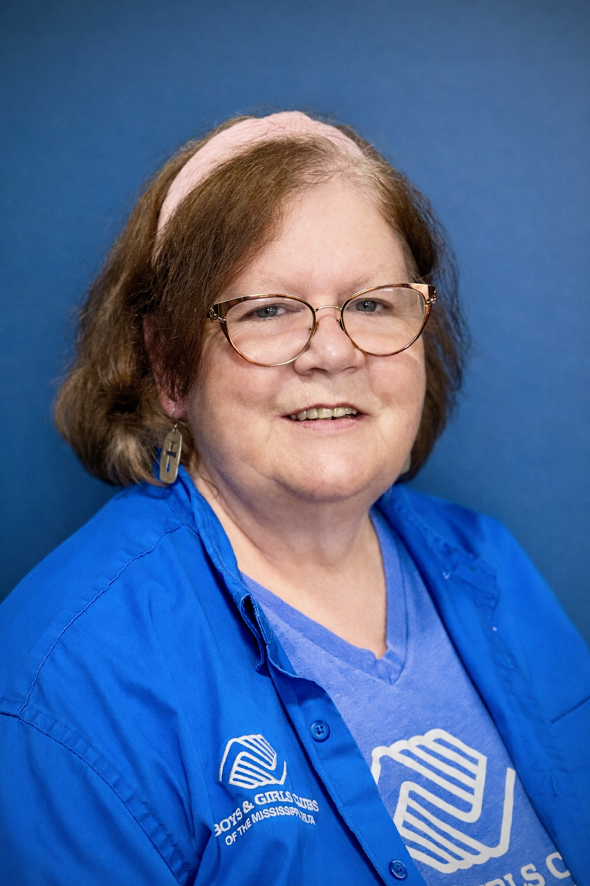 A woman with short brown hair, glasses, and a pink headband, smiling, wearing a blue shirt and a blue jacket with the logo of the Boy Scouts of America on it, standing against a blue background.