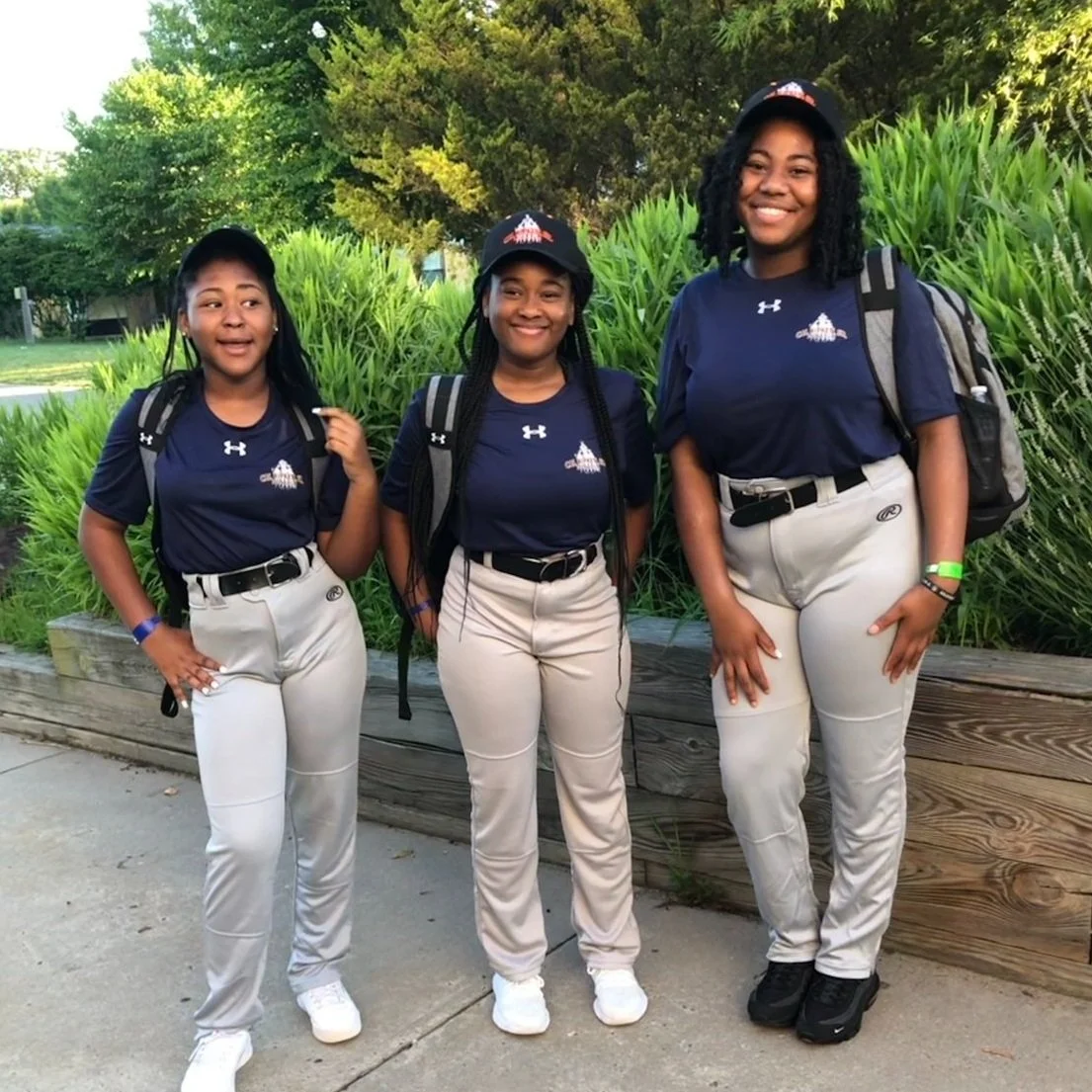 Three young girls in matching blue sports shirts and beige baseball pants standing outdoors in front of green grass and trees, smiling at the camera, each wearing a black cap and carrying backpacks.