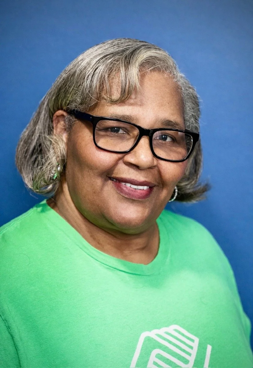 A woman with gray hair and glasses smiling, wearing a green shirt with a Boys & Girls Clubs logo, standing against a blue background.