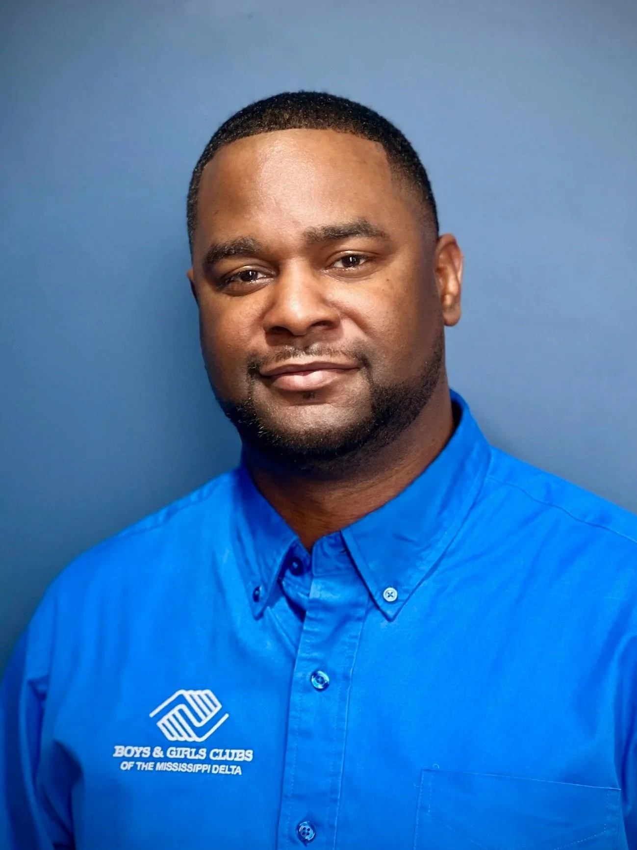 A man with short black hair and a beard wearing a blue button-up shirt with the Boys & Girls Clubs logo on it, standing against a blue background.