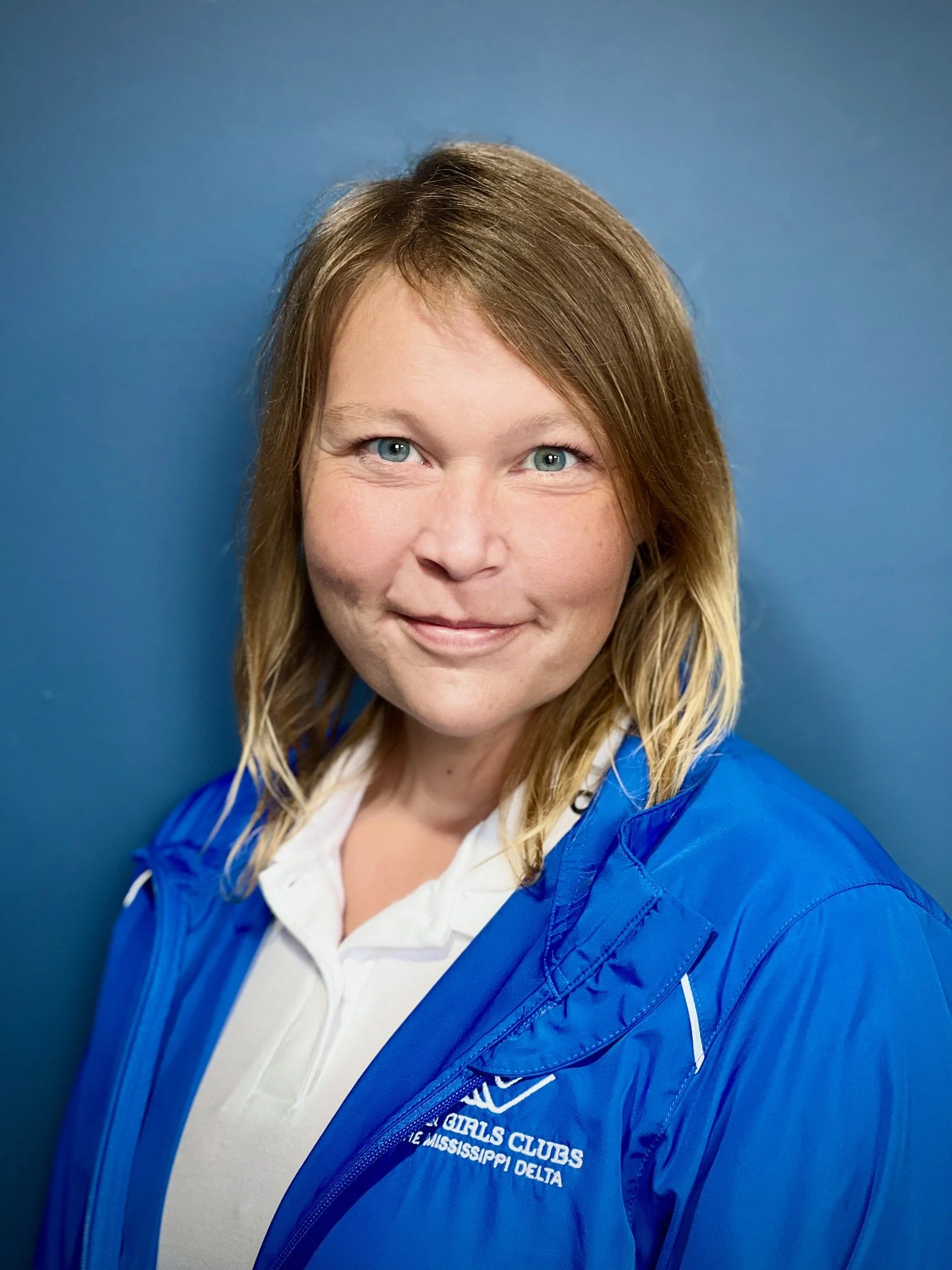 Portrait of a woman with shoulder-length light brown hair, blue eyes, and fair skin, wearing a white polo shirt and a blue jacket with a Girls Clubs logo, standing against a blue background.