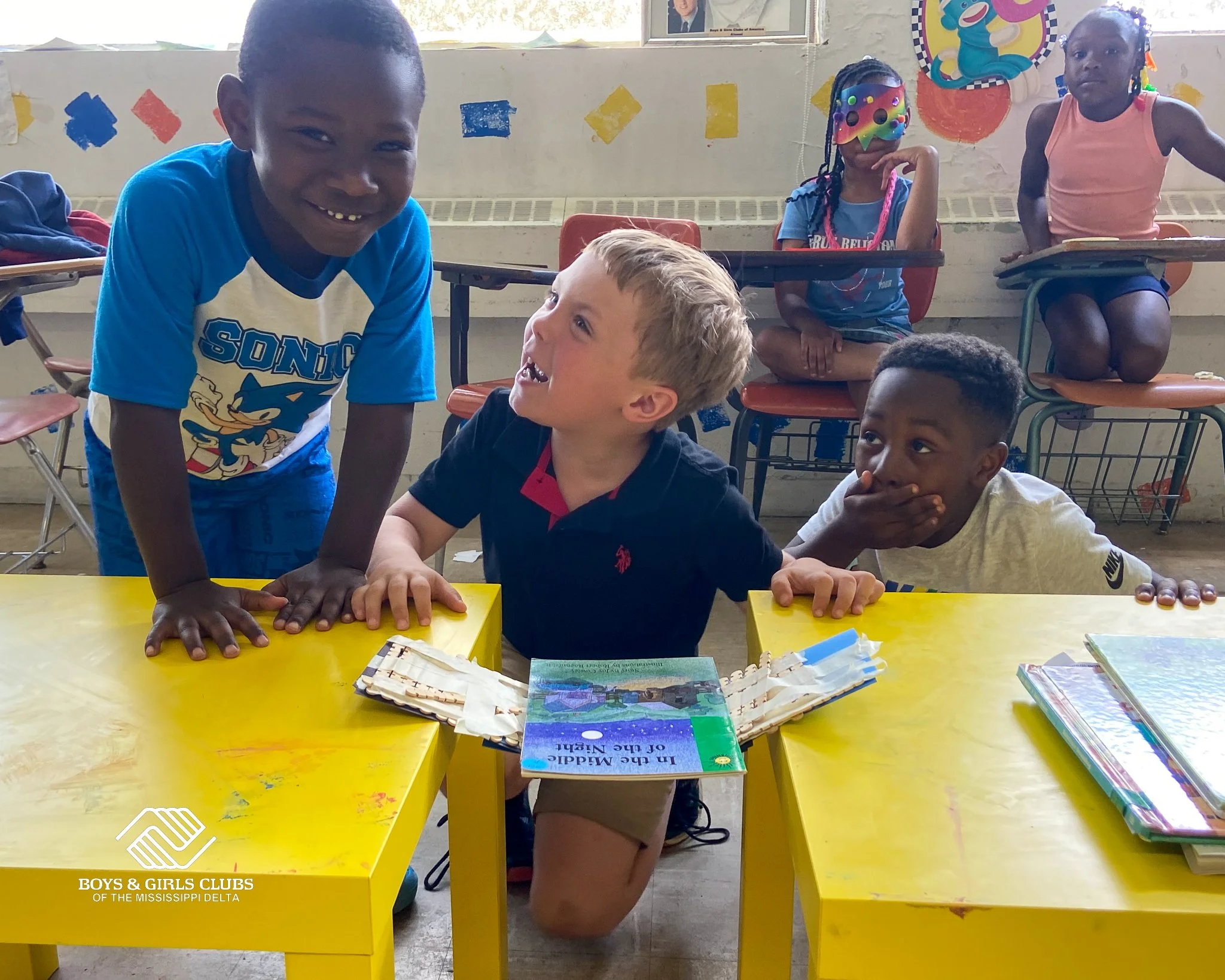 A classroom scene with children. A boy in a blue Sonic shirt smiles towards the camera.  girl with braces and a black shirt is talking to a blonde boy sitting at a yellow table, with books and papers in front. Two other girls are seated on chairs, on