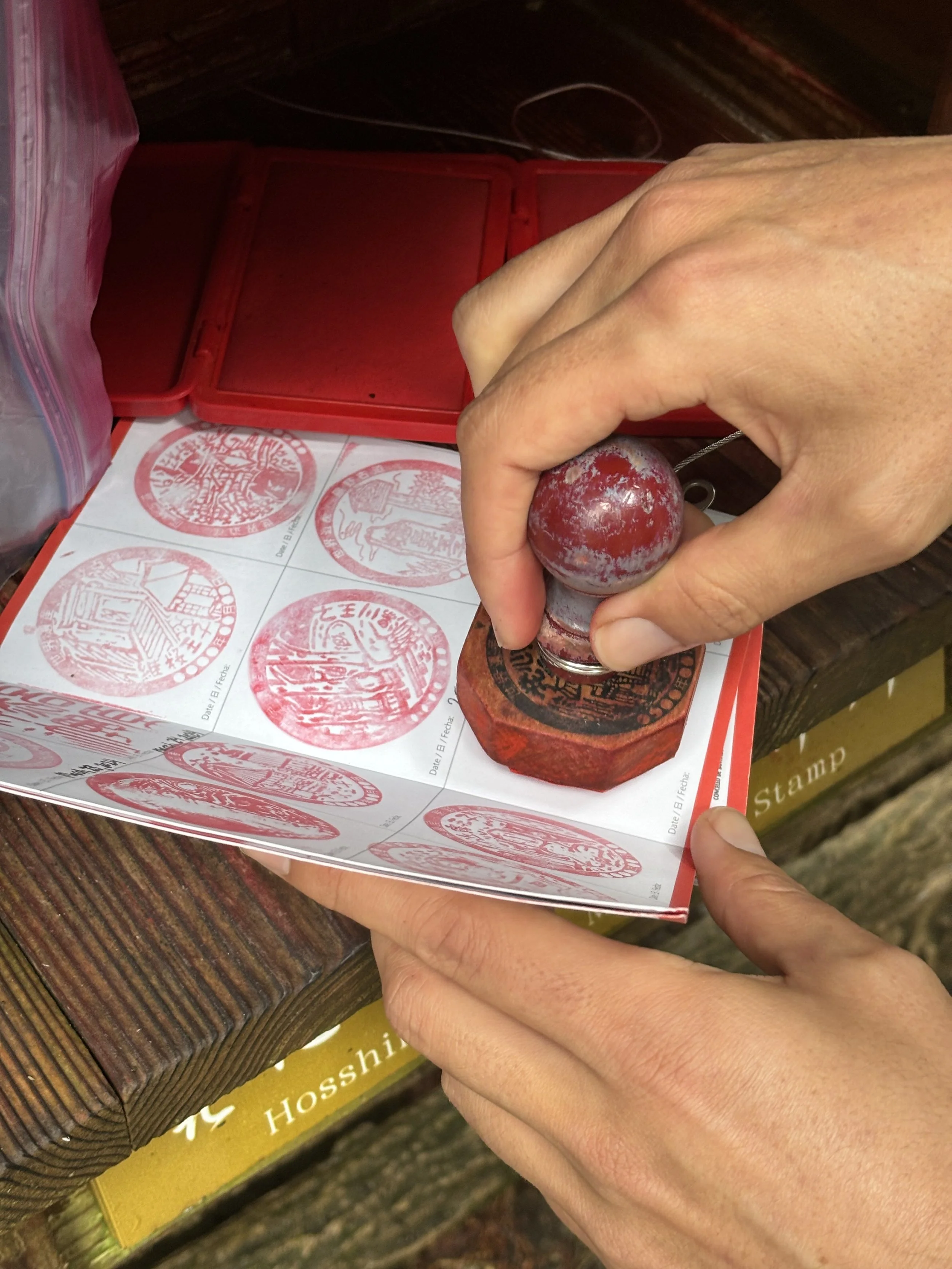 Close-up of a person stamping a red circular ink stamp onto paper using a carved wooden stamp, with more stamped papers visible in a red box.