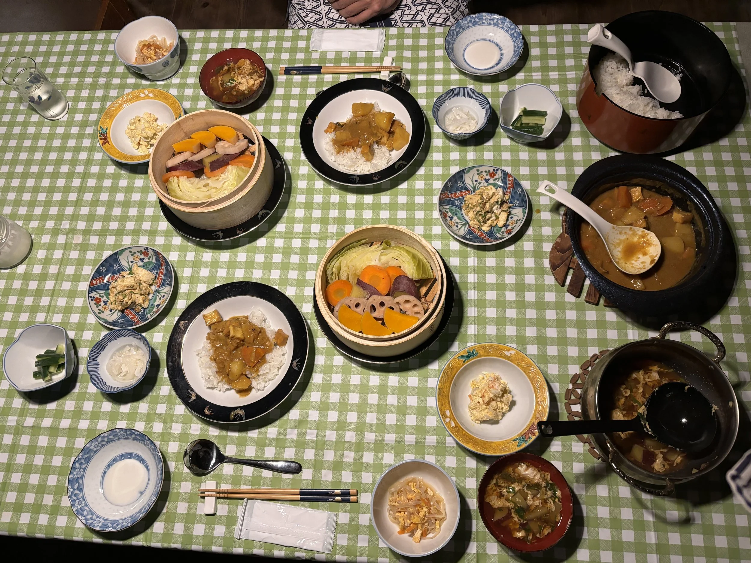 A table set with various dishes of Japanese cuisine, including bowls of rice, curry, and vegetables, along with small side dishes, chopsticks, and serving spoons on a green checkered tablecloth.
