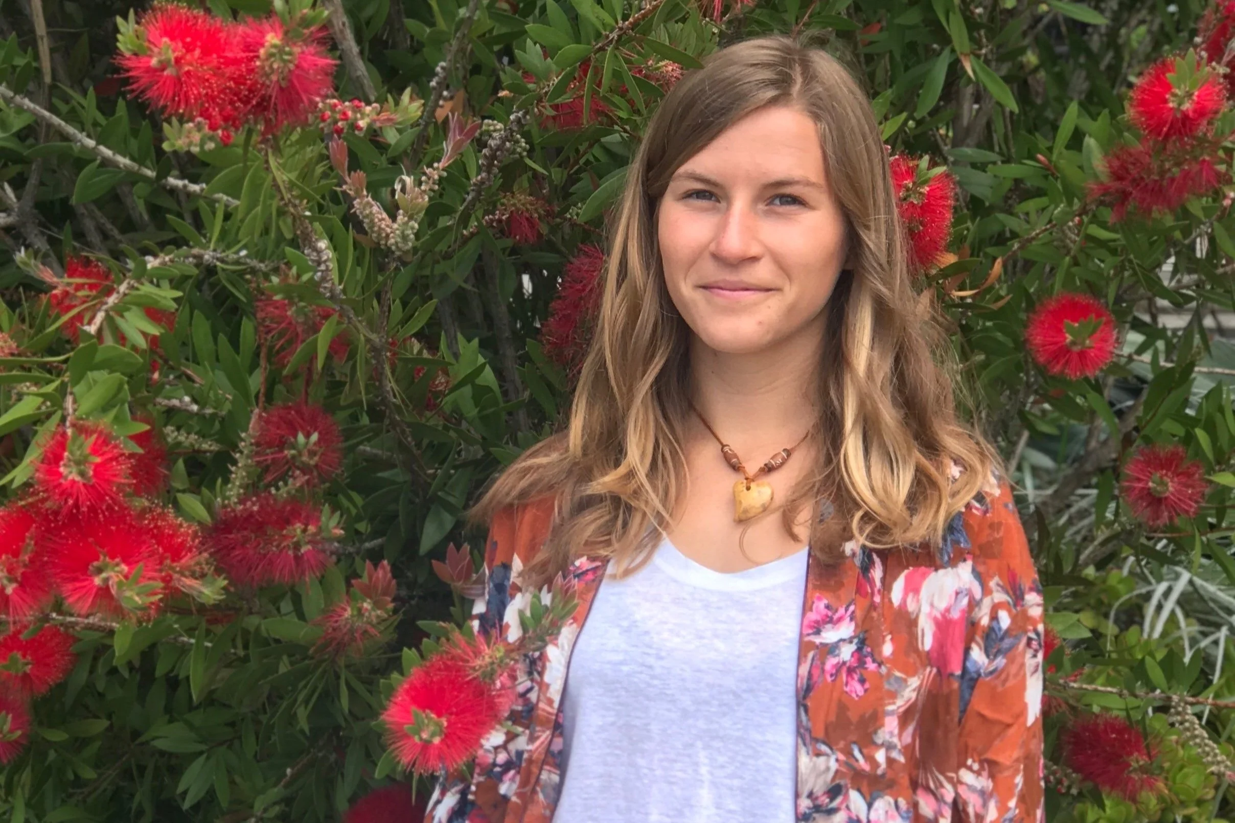A young woman with light brown, curly hair standing in front of a bush with red, fluffy flowers and green leaves, wearing a white shirt, a colorful floral jacket, and a tan heart-shaped necklace.