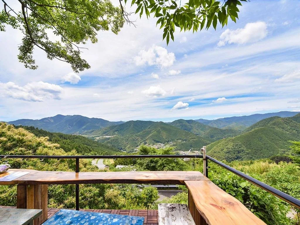 A scenic view of green mountains and a partly cloudy sky from a balcony with a wooden railing and a wooden table.