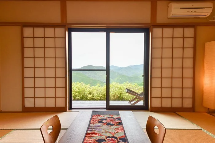A traditional Japanese room with tatami mats, shoji sliding doors opening to a balcony with a mountain view, and a low wooden table with a decorative runner.