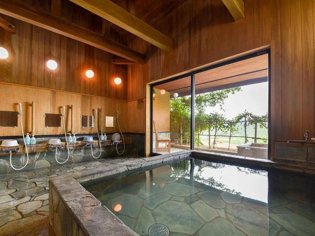 Interior of a traditional Japanese-style onsen with a stone hot spring bath, wooden walls, and shower stations with handheld showers and soap bottles, overlooking a garden through a large sliding glass door.