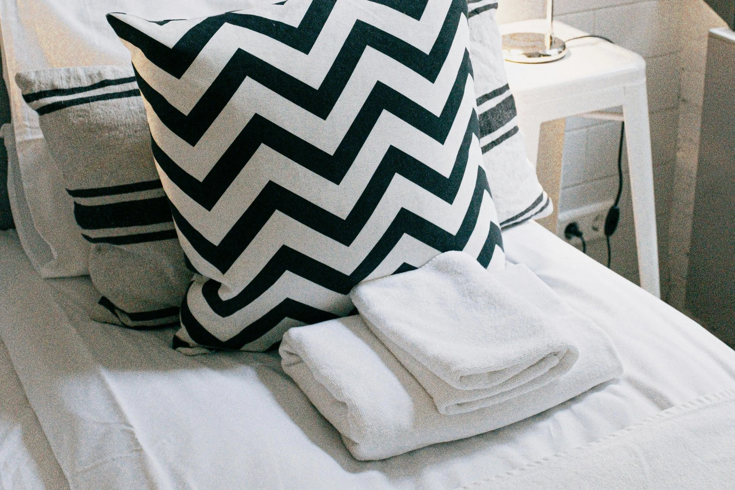A neatly made bed with black and white zigzag-patterned pillows, white towels folded on top, and a bedside table with a lamp.