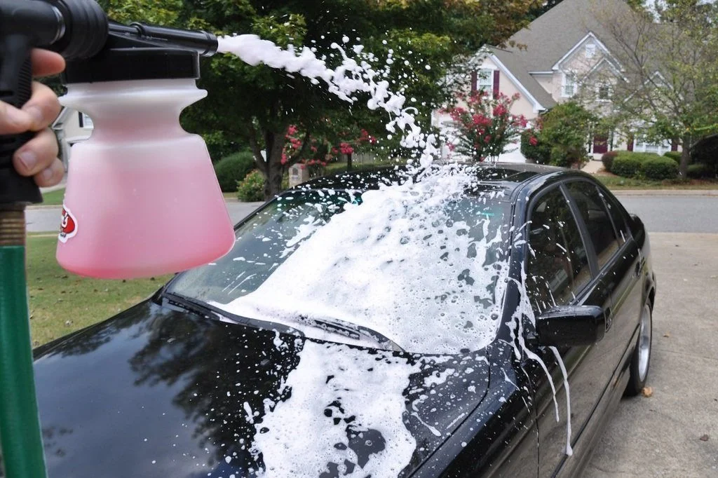 A person using a foam cannon to wash a black car outdoors, with foam splashing onto the windshield and hood, in a neighborhood setting.
