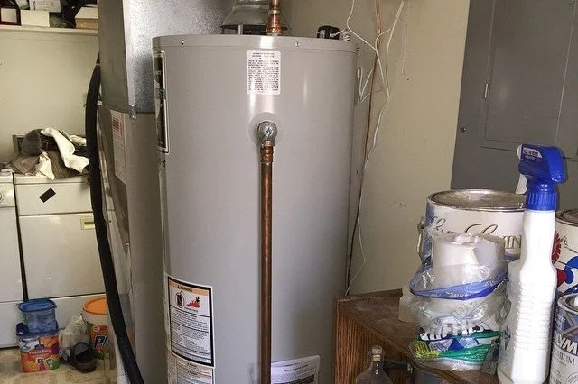 A water heater in a utility room with various household items around it, including a cleaning spray bottle and plastic containers.