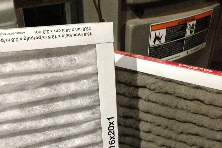 Two air filters placed on a surface, one new and unused with white pleats, the other used with grayish pleats.