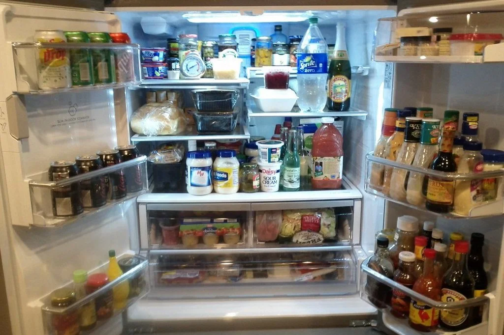 Open refrigerator with various food and beverage items including condiments, jars, sauces, drinks, and fresh produce.
