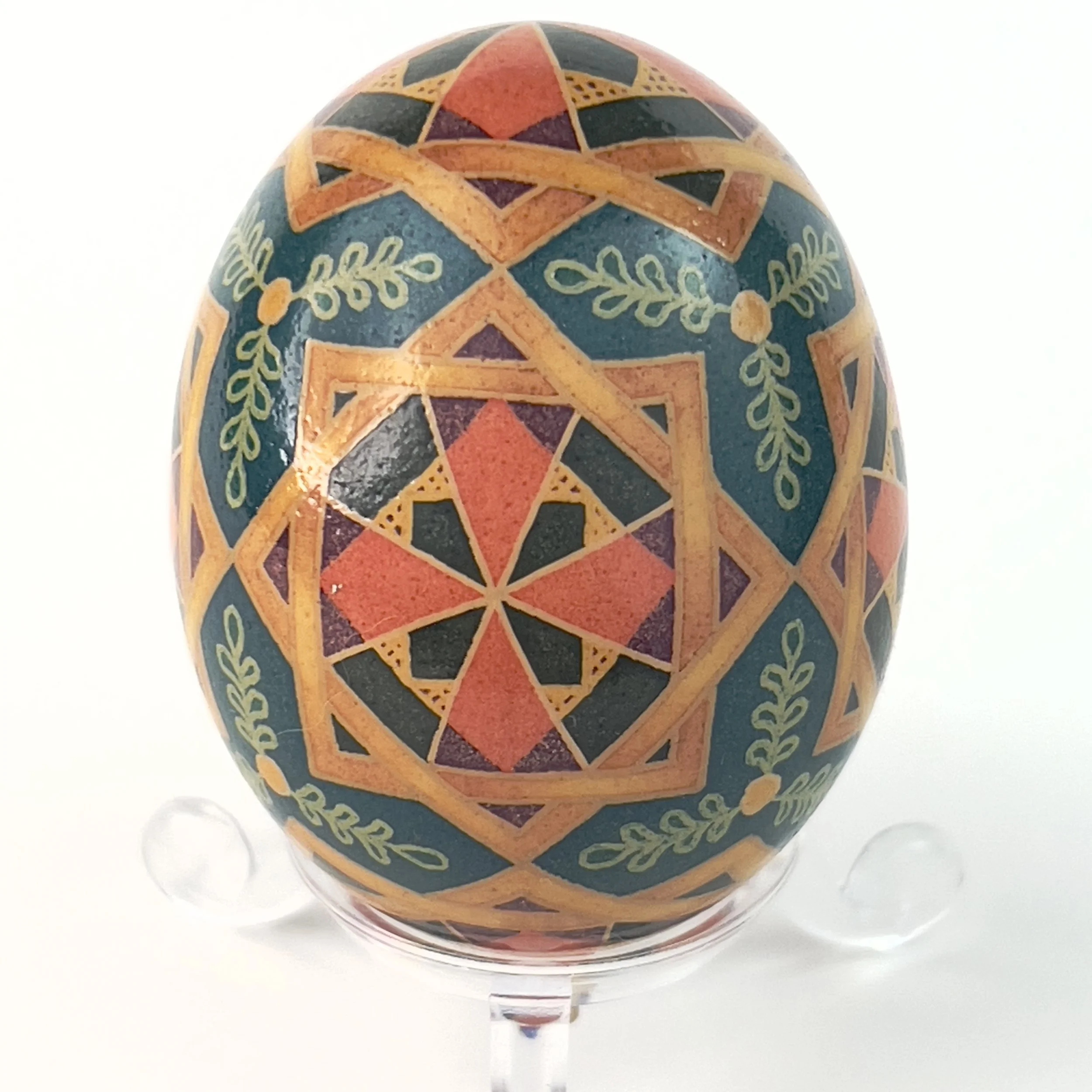 Decorated egg with geometric and floral patterns in orange, blue, red, and green, displayed on a clear stand against a white background.