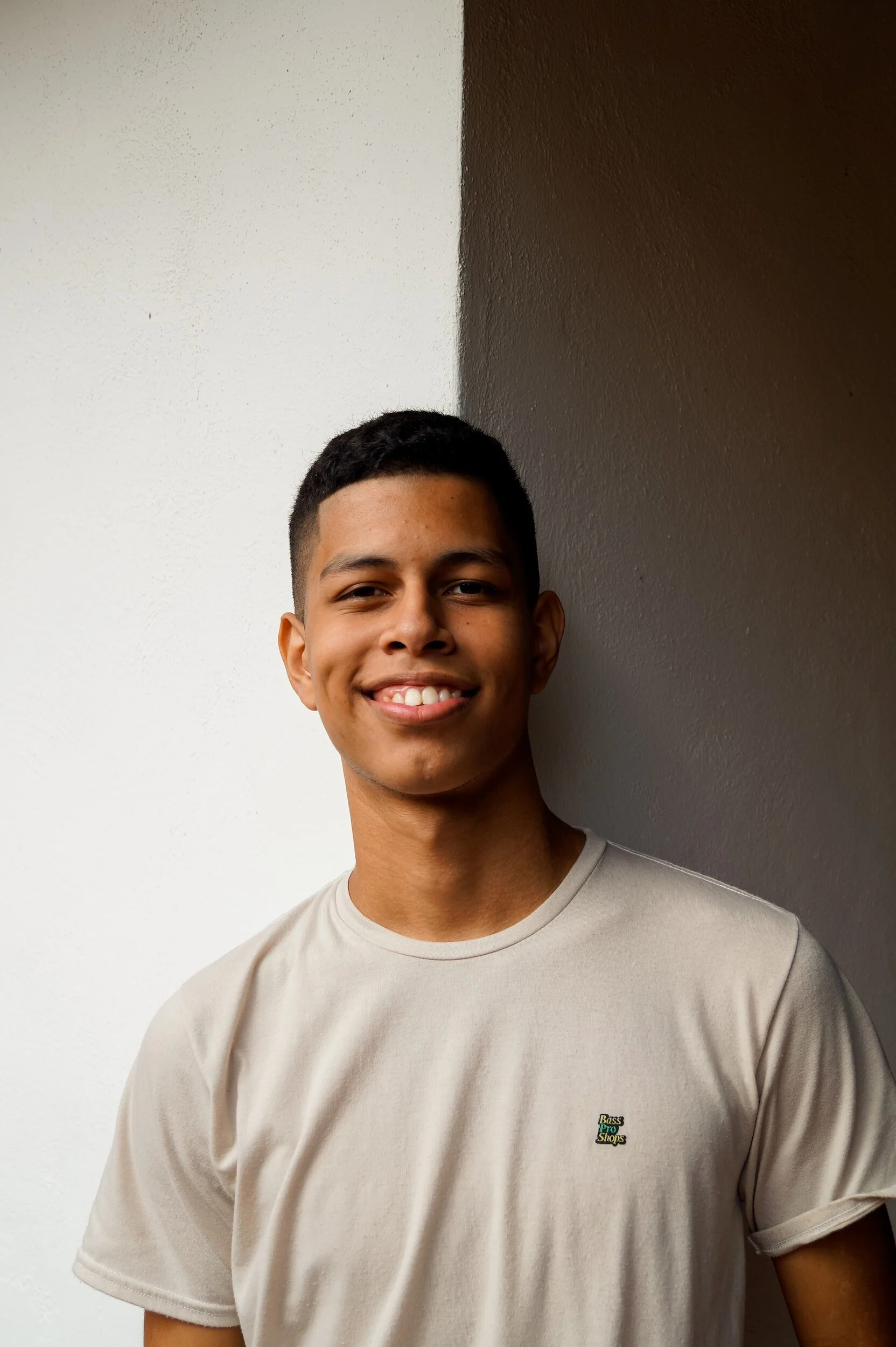 Smiling young man wearing a beige t-shirt, standing against a white and brown wall.