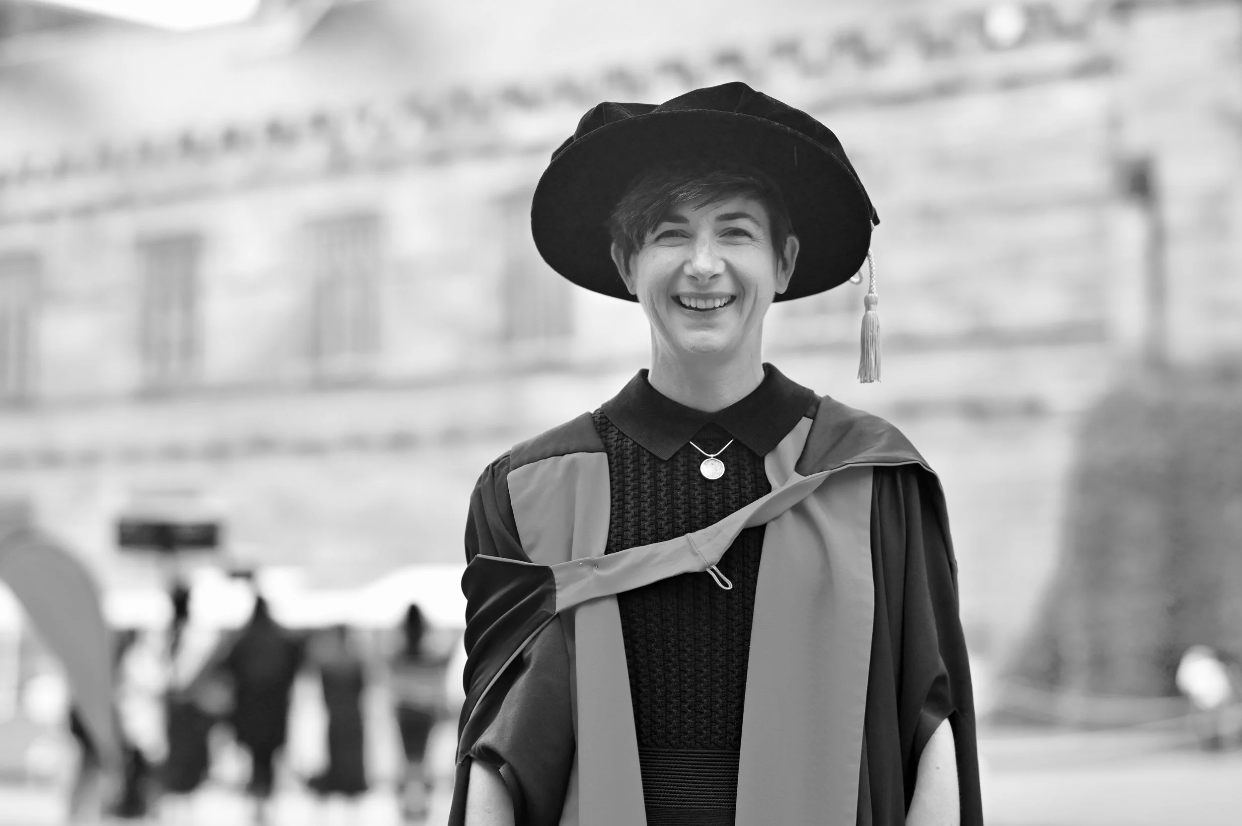 A young woman in graduation attire, smiling and wearing a cap with a tassel, standing outdoors with a blurred background.