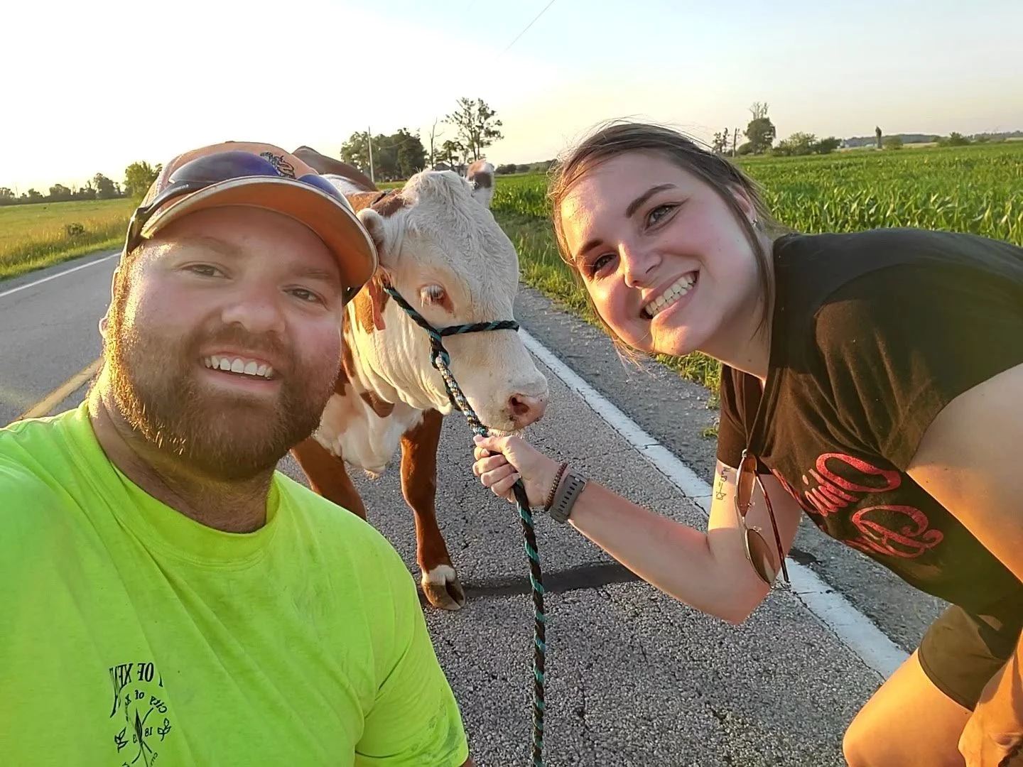 Two smiling people, a man wearing a yellow shirt and a woman wearing a black shirt, are posing next to a cow on a sunny rural road, with the cow's head gently touching the woman's hand.