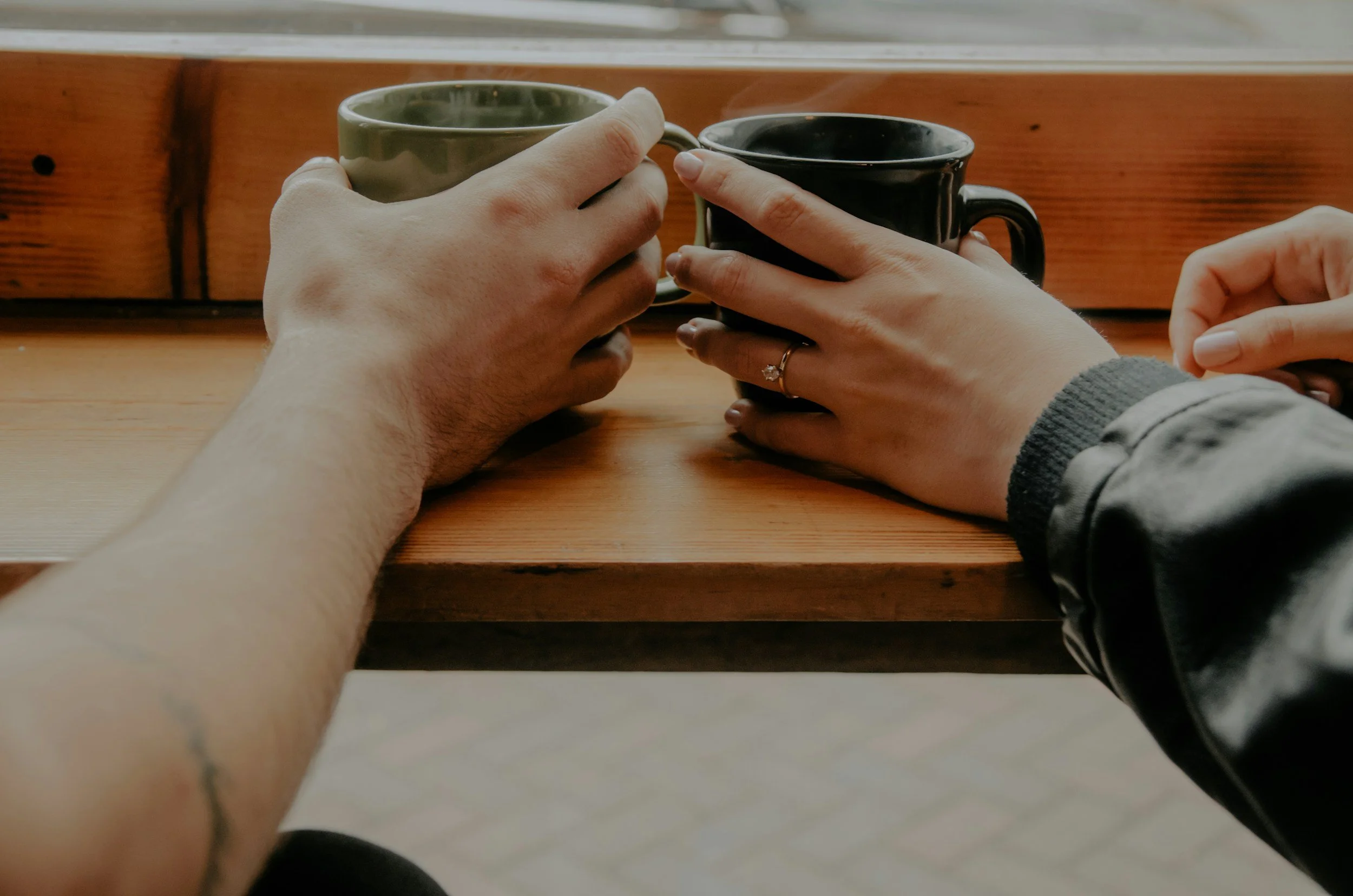Two people holding black and green coffee mugs at a wooden table.