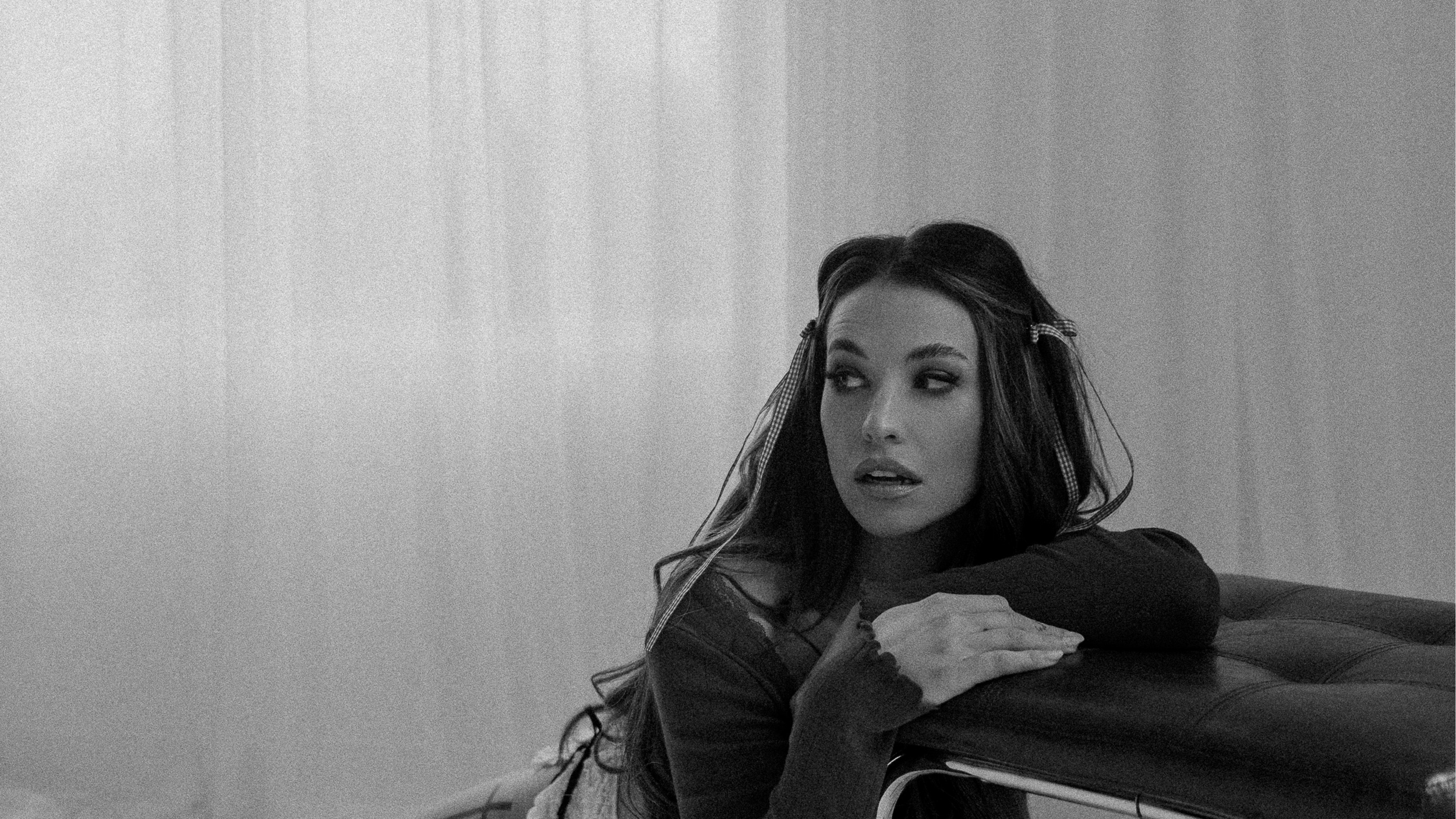 A young woman with long wavy hair and hair clips is resting her chin and arms on a cushioned bench, looking to the side with a thoughtful expression.