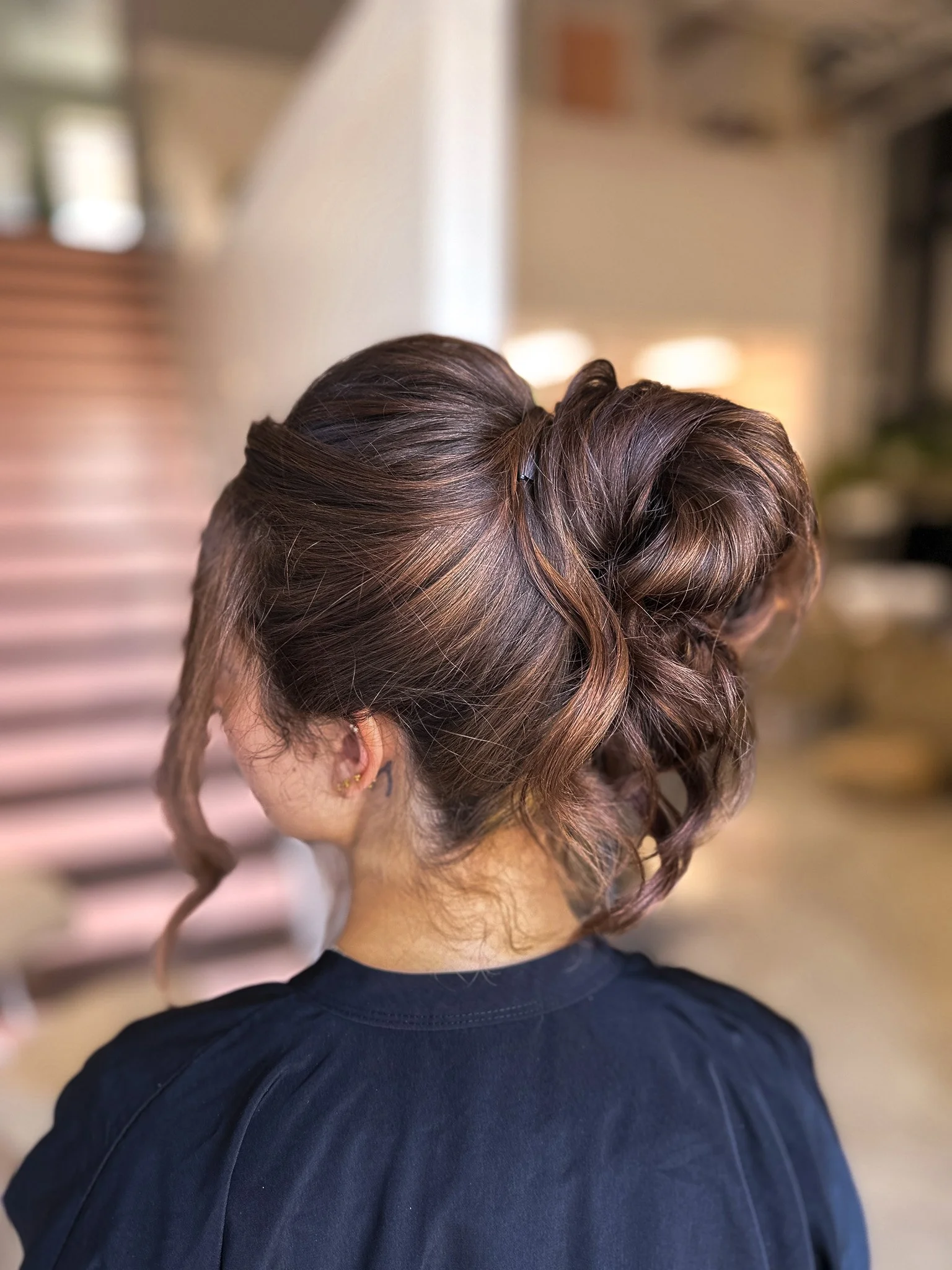 Back view of a woman with a styled updo hairstyle featuring loose curls, wearing a black top, in an indoor setting with blurred background.