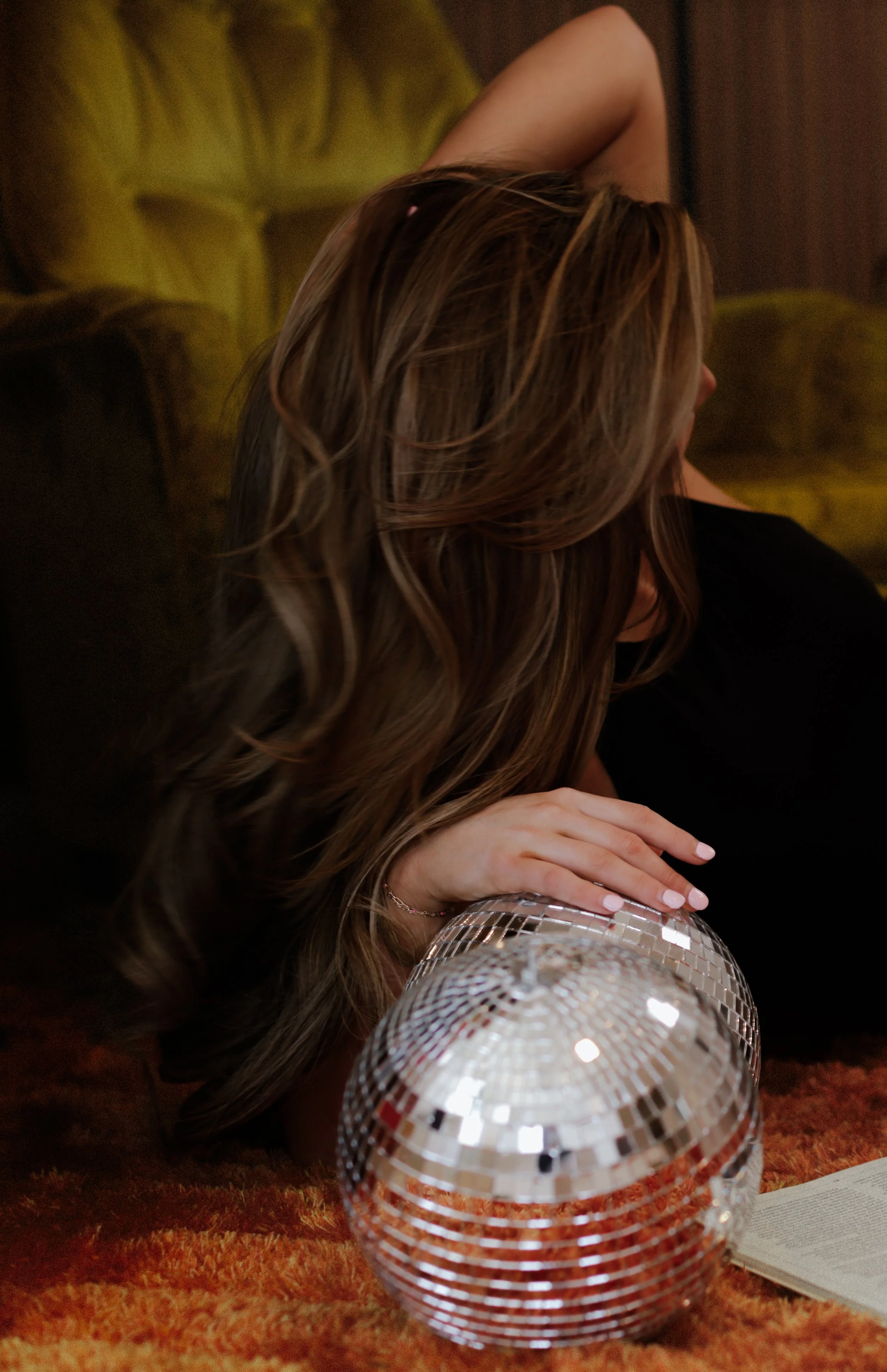A woman with long, wavy hair lying on her side on an orange textured carpet, propped up on one elbow with her hand resting on a disco ball. She is wearing a black top. There is an open book nearby, and a green armchair is in the background.