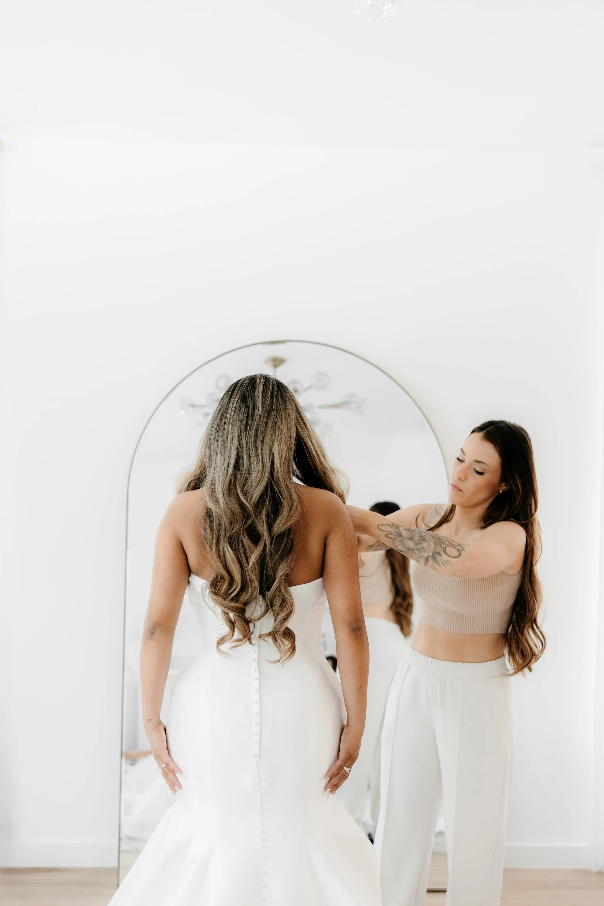 A woman in a wedding dress is getting assistance from another woman in casual attire as she prepares for her wedding.