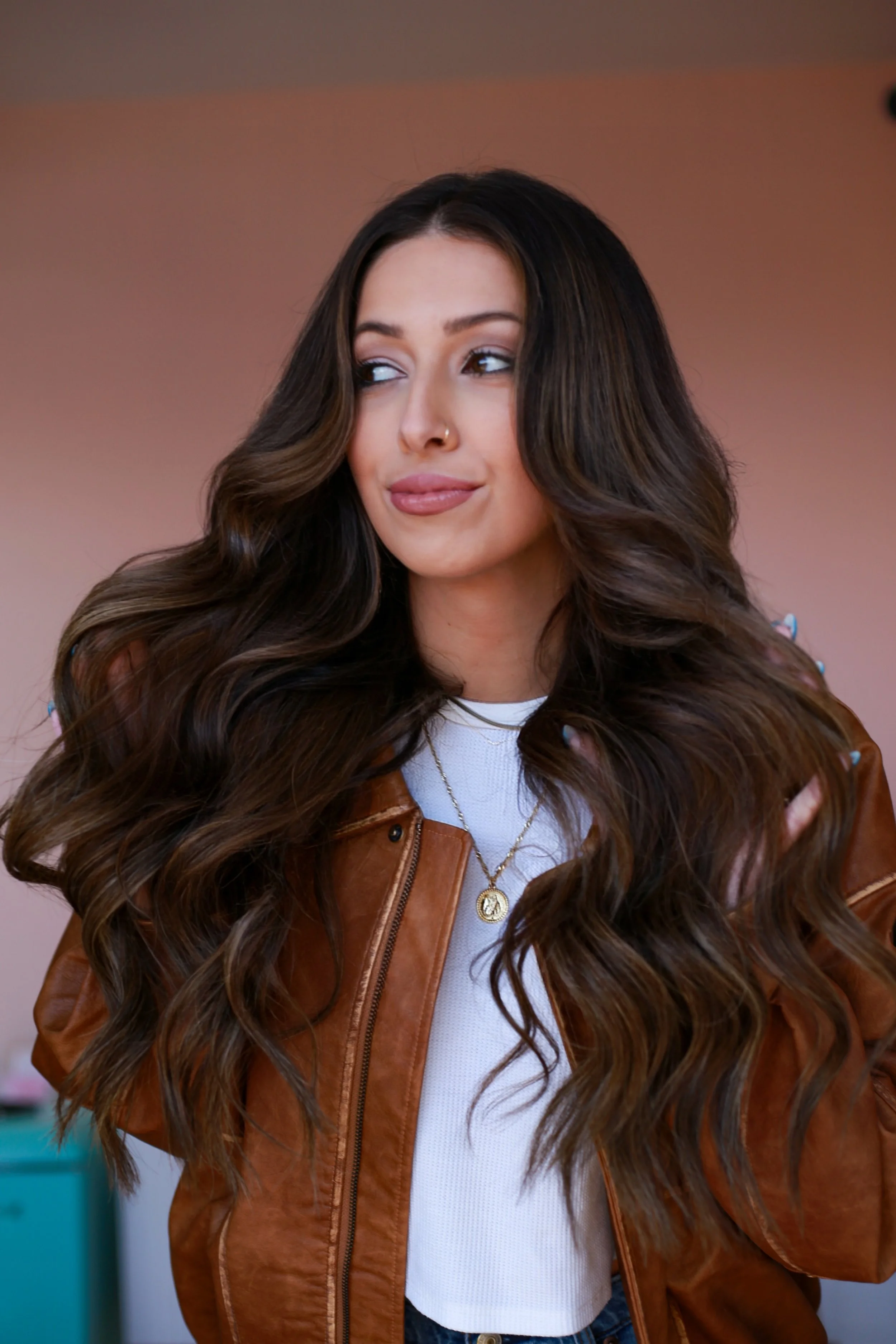 A woman with long wavy brown hair, wearing a brown leather jacket, white shirt, and a gold necklace, posing indoors with a pinkish background.