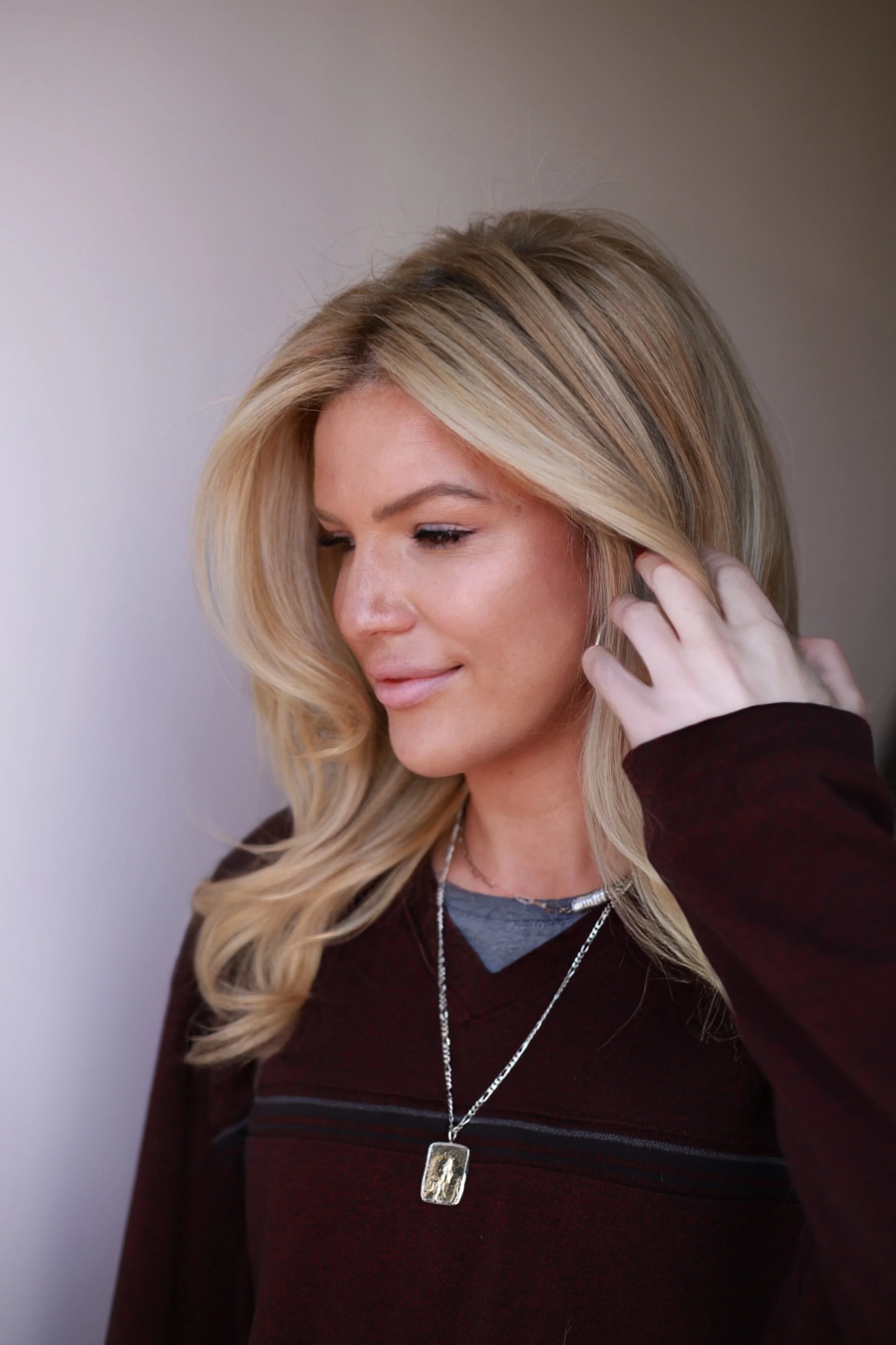 A woman with blonde hair, wearing a maroon top and a silver necklace, touching her hair and smiling softly.