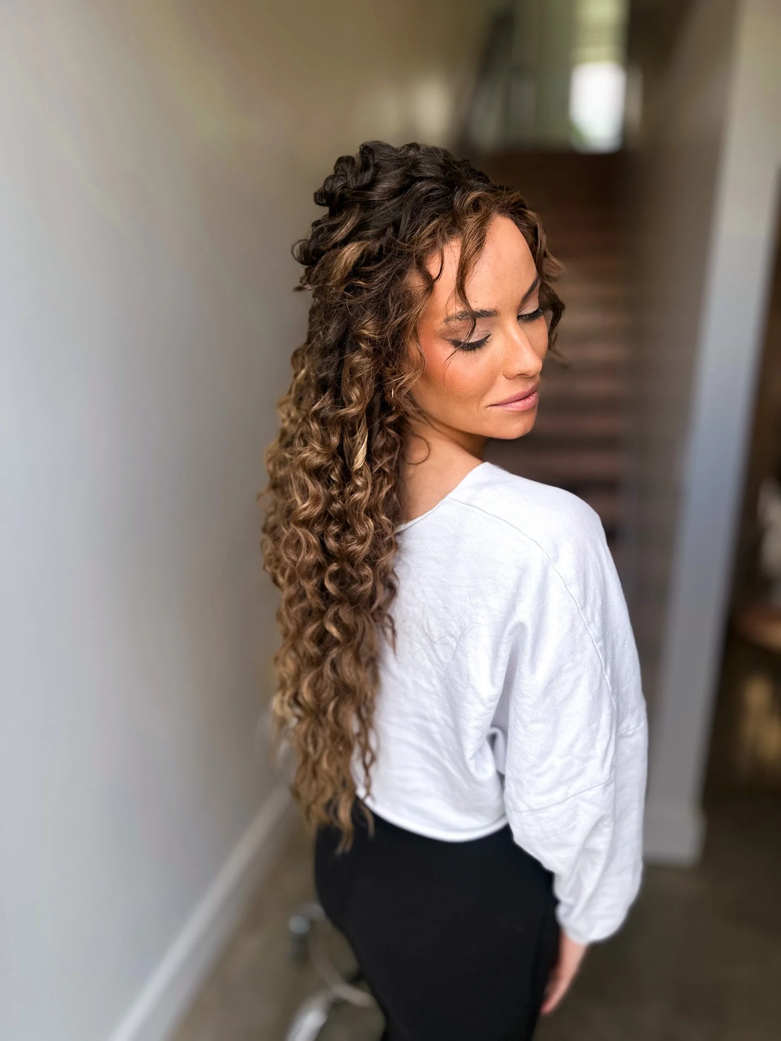 A woman with long, curly brown hair, wearing a white top, standing in a hallway.