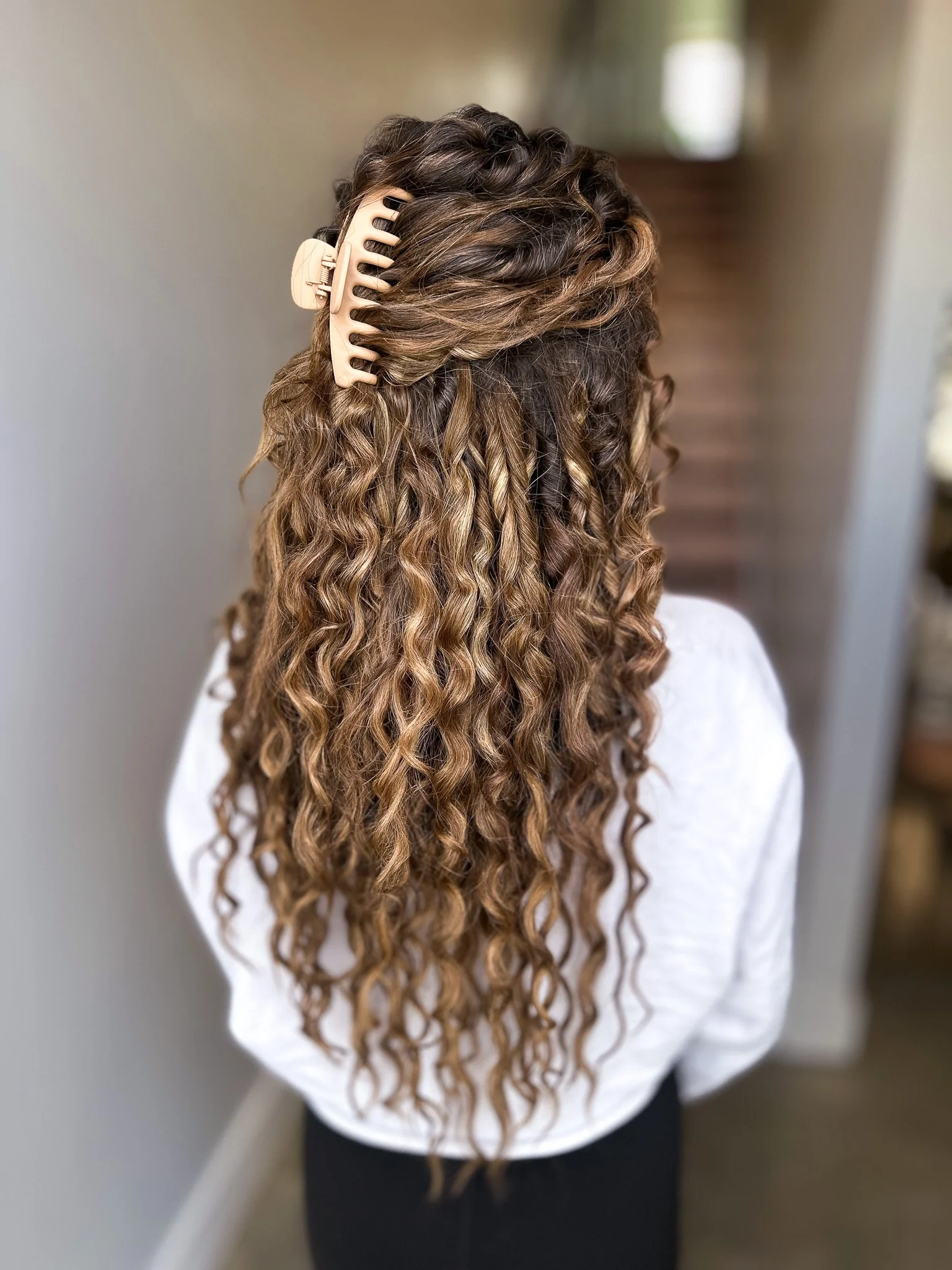 A person with long, curly, light brown hair secured with a beige hair clip, standing indoors facing away from the camera.