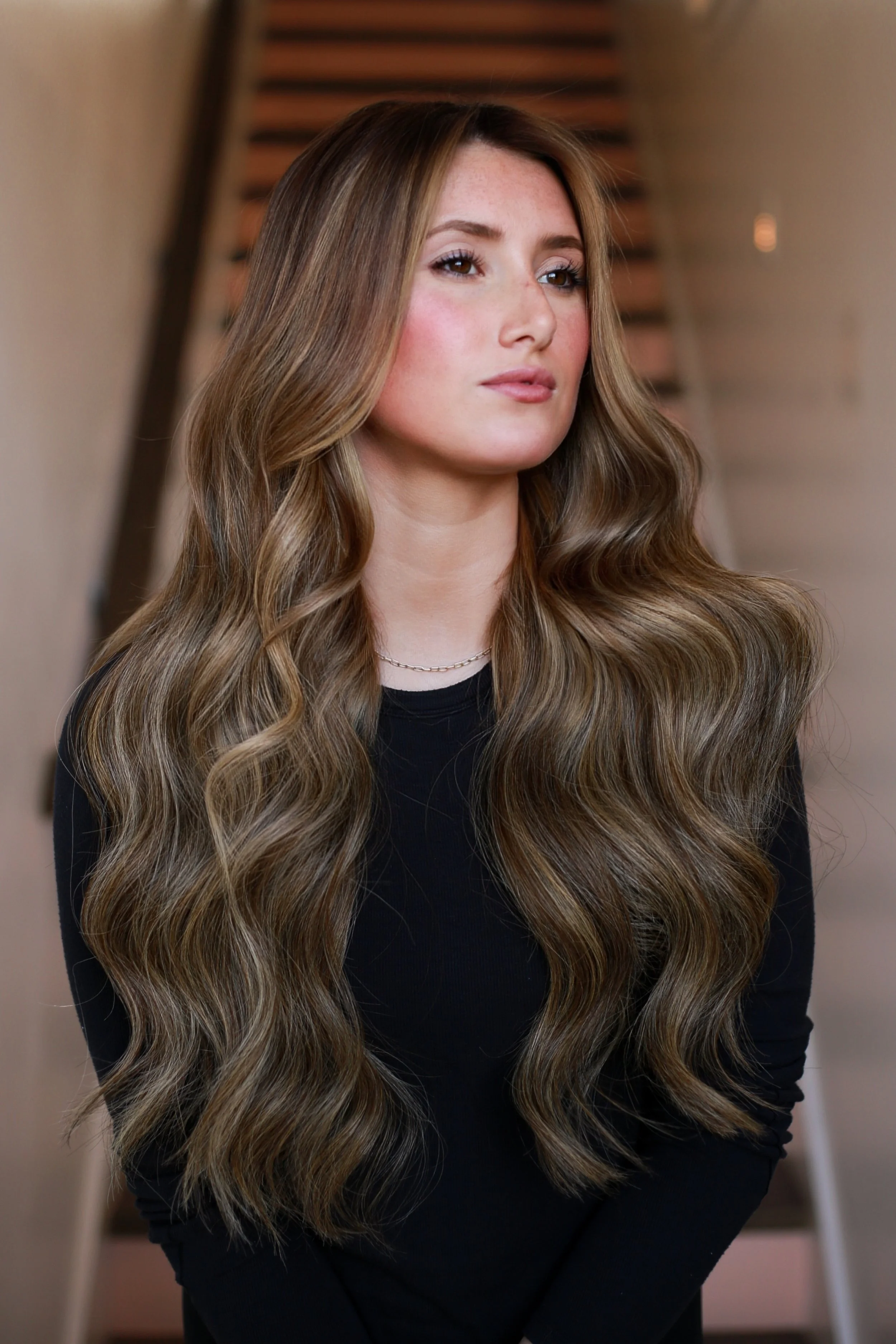 Young woman with long, wavy brown hair and a black top standing indoors in front of a staircase.
