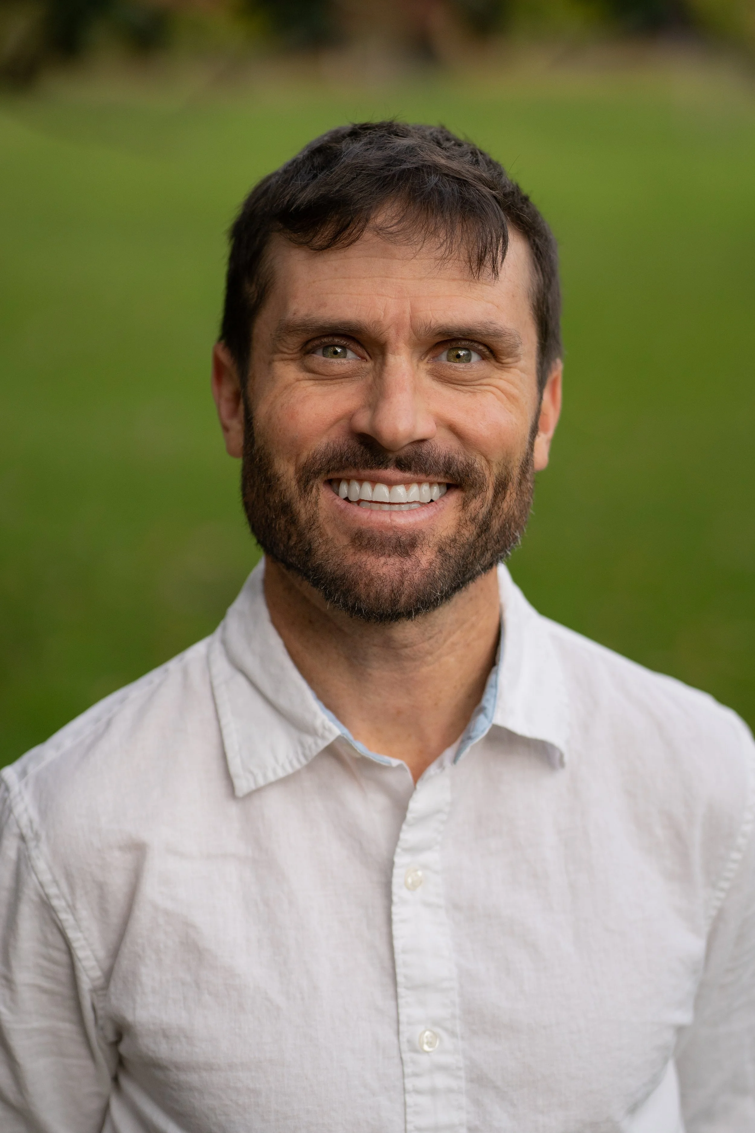 A man smiling outdoors, wearing a white shirt, with a blurred green background.