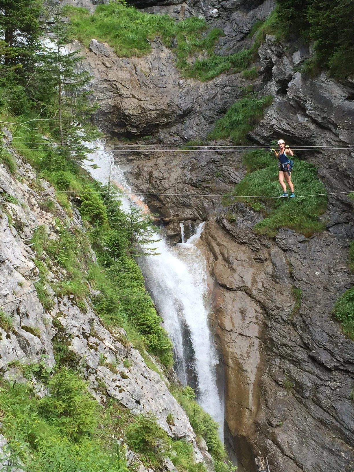 Active individual walking on a tightrope across a mountain landscape with a waterfall below and green trees surrounding the area.