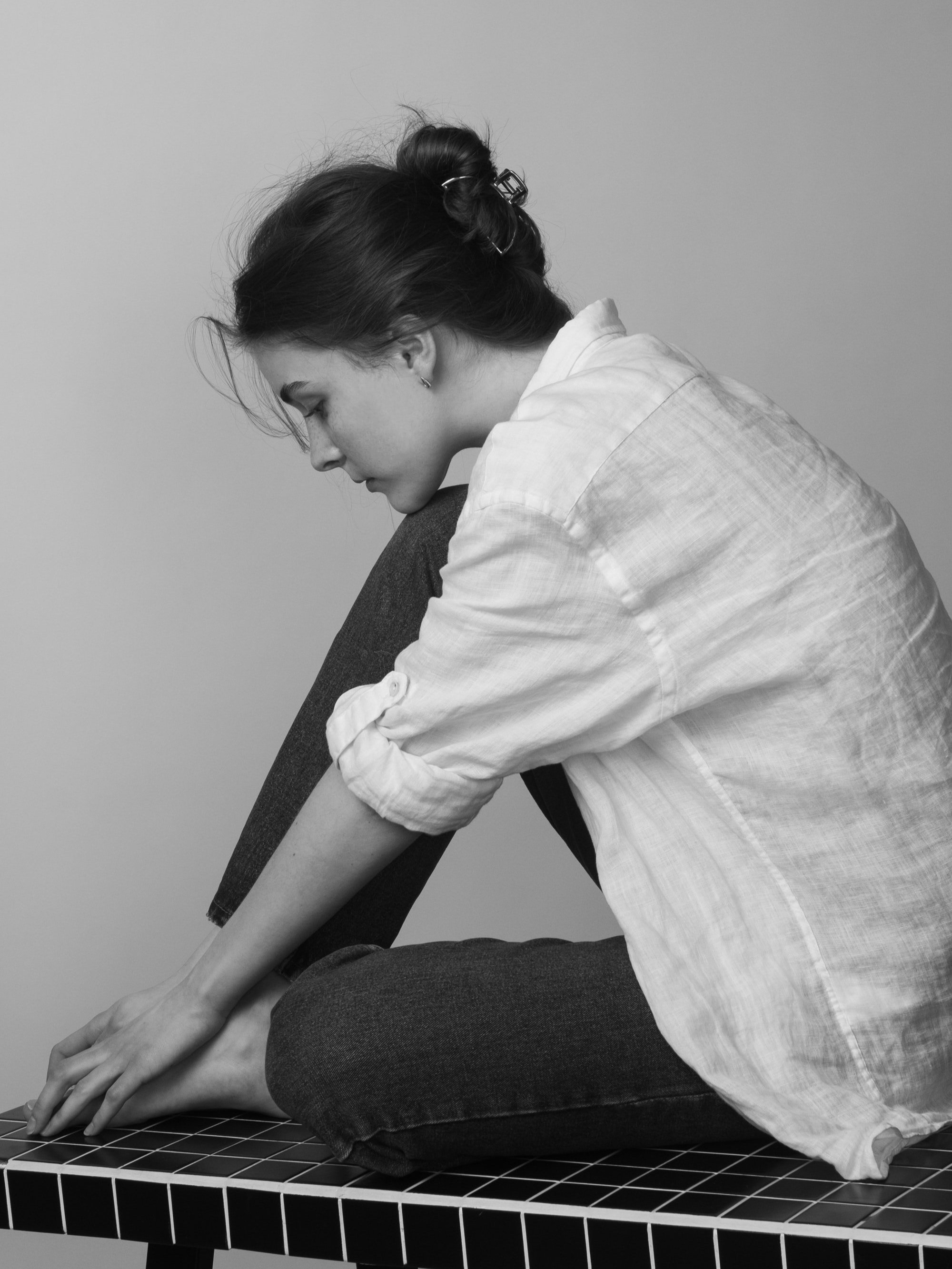 A woman with dark hair tied up, sitting in a cross-legged position on a tiled surface, with her head bent down and her hands reaching toward her toes.
