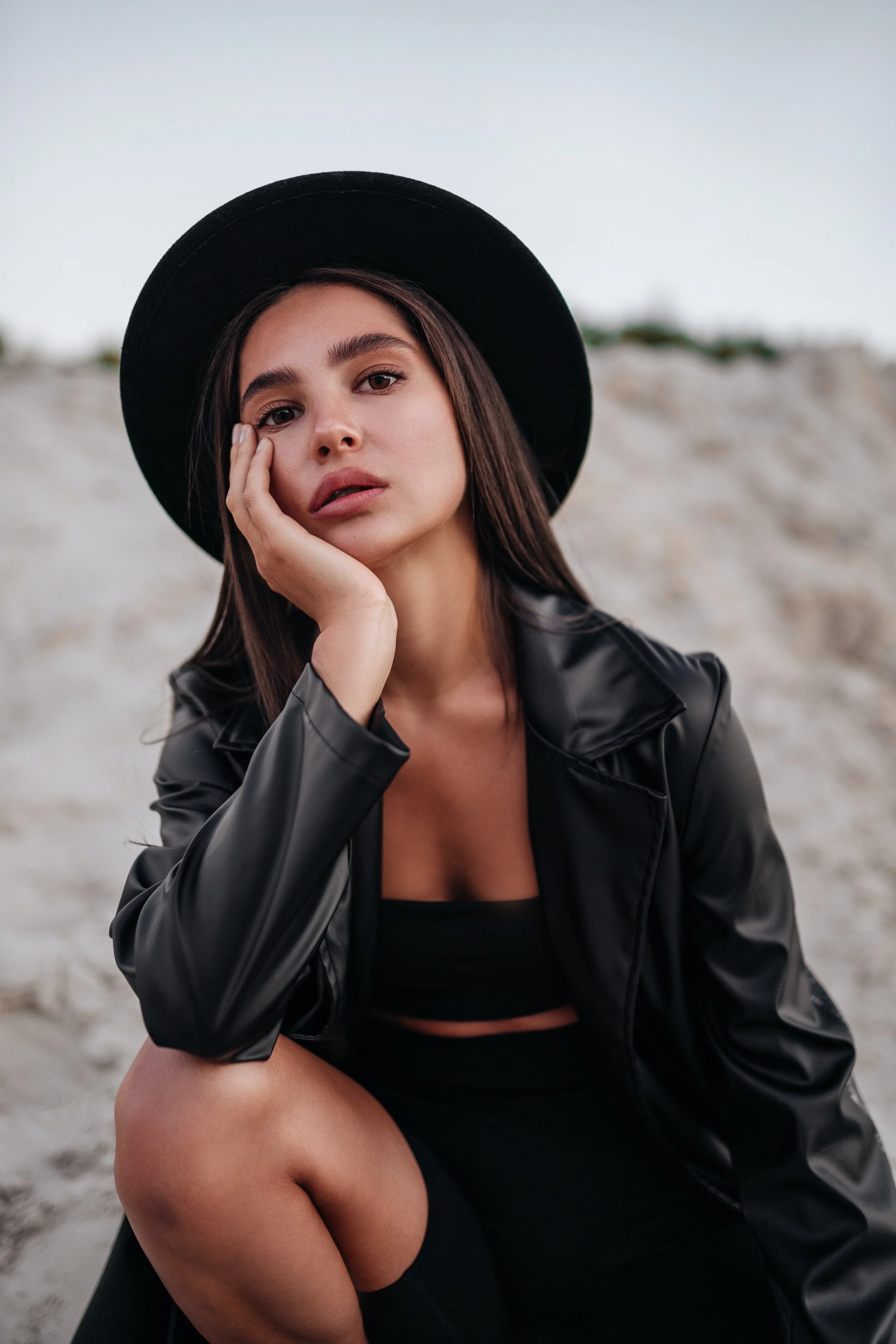 A young woman with long brown hair, wearing a black wide-brim hat, black leather jacket, and black crop top, sitting on sand with her hand resting on her face, outdoors with cloudy sky.
