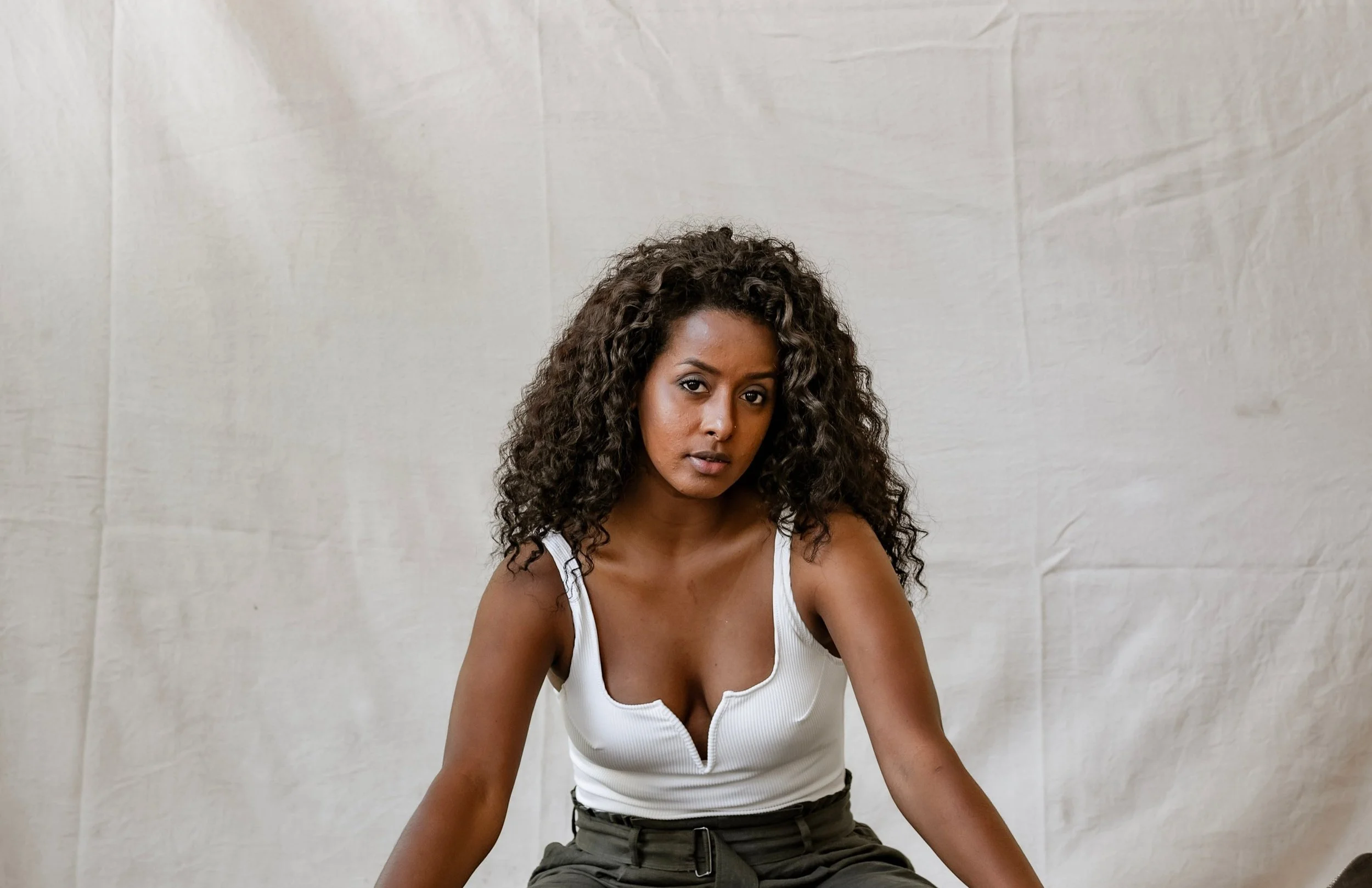 A woman with curly hair wearing a white tank top and gray pants sitting against a light background.