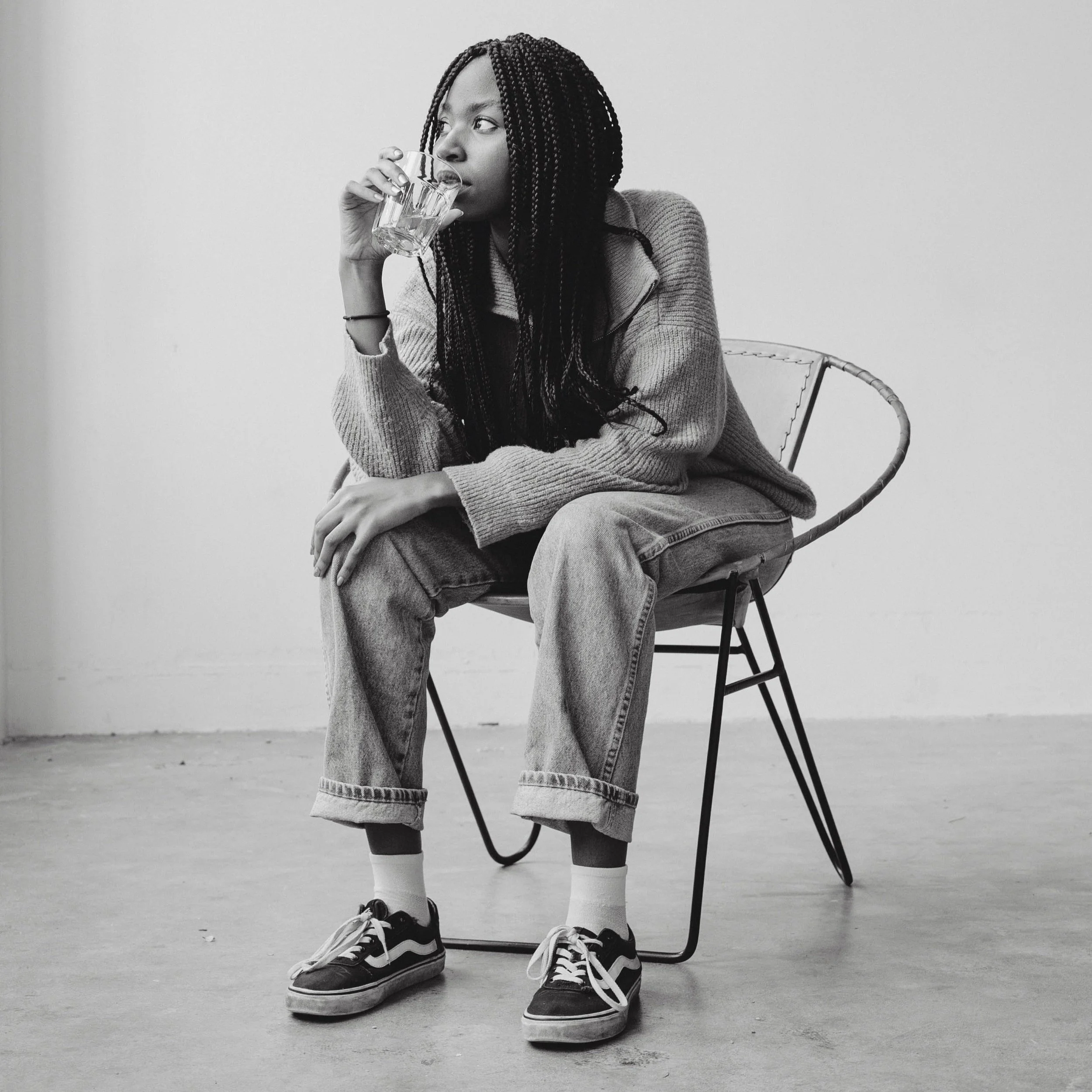 A young woman with braided hair sitting on a modern chair in a room with a plain wall, drinking water from a glass.