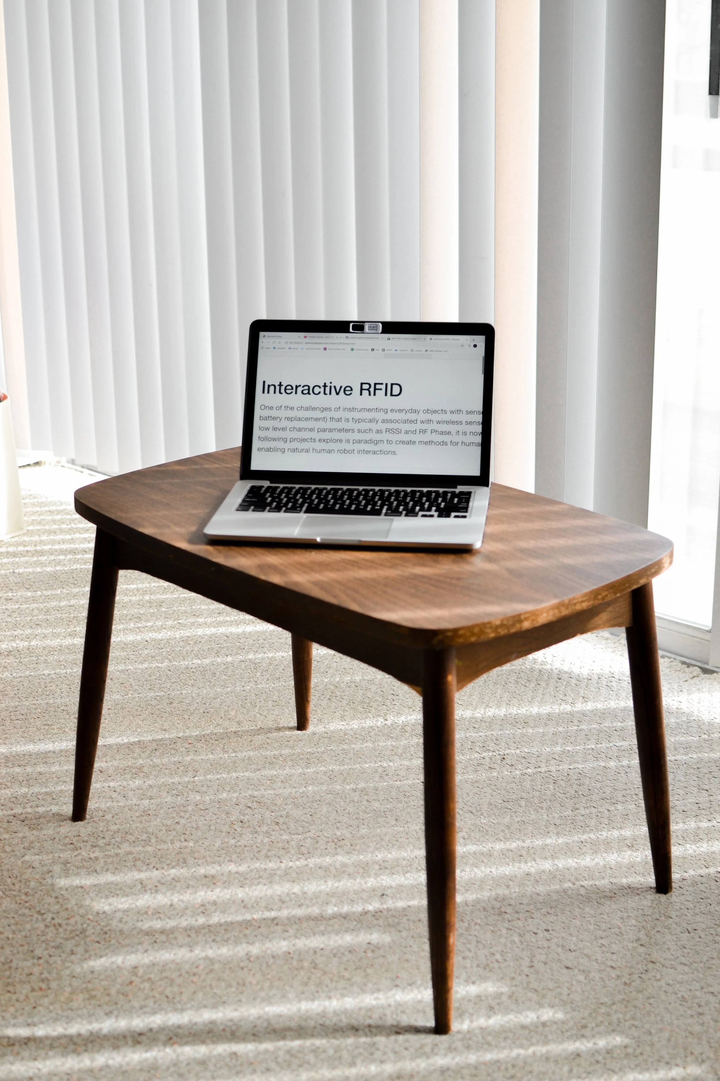 A wooden table with a laptop displaying the text 'Interactive RFID' in a room with vertical blinds and beige carpet.