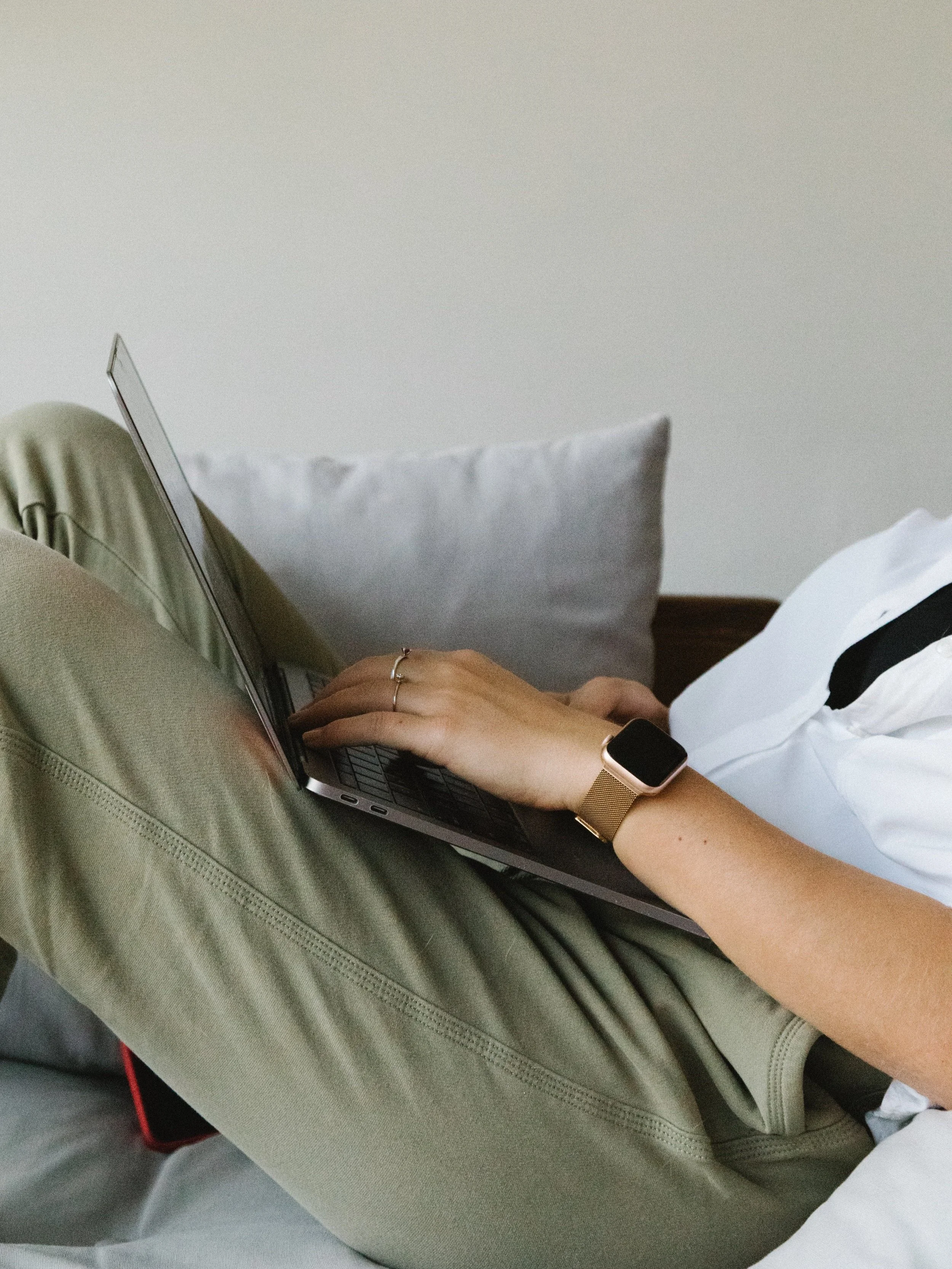 Person lying on bed using a laptop, wearing an Apple smartwatch and a ring, with a gray pillow in the background.