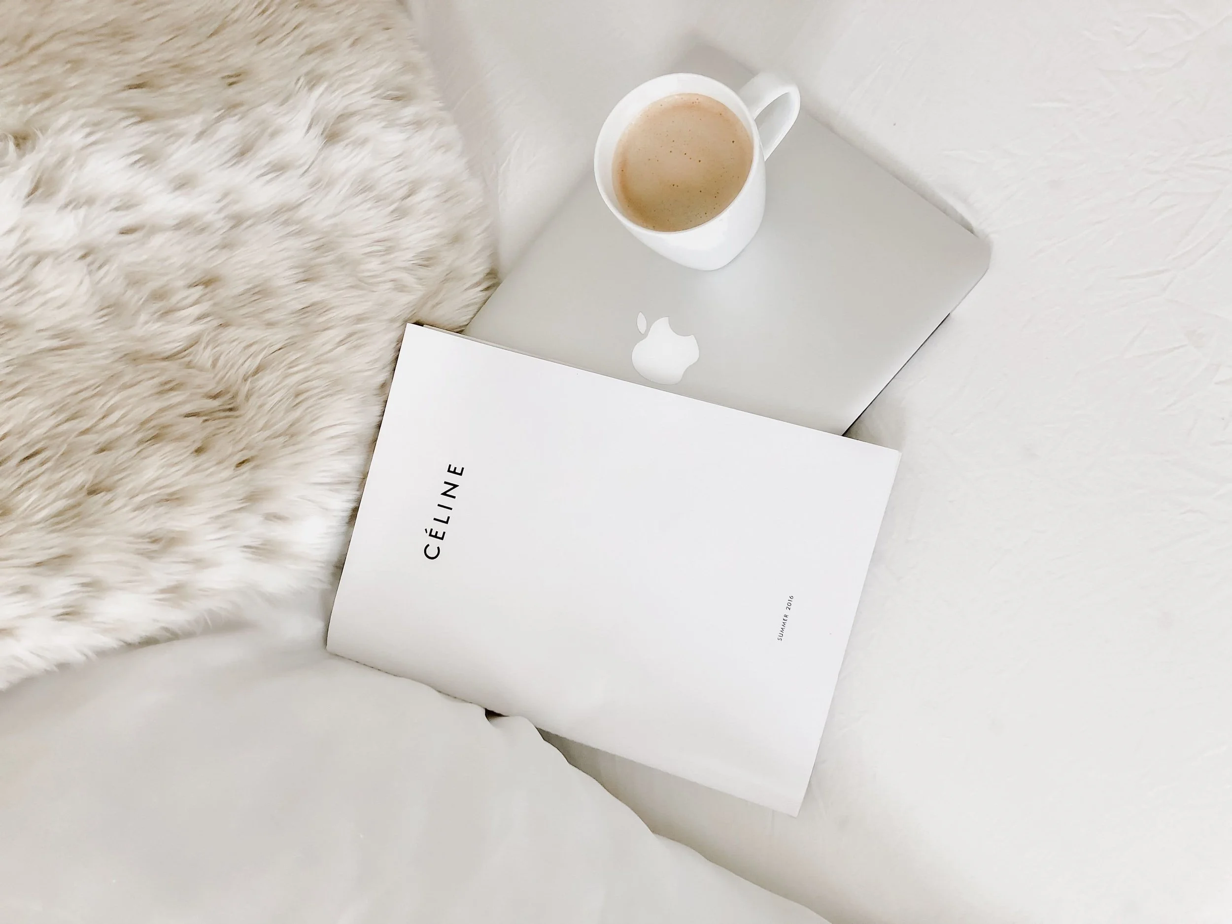 A flat lay of a white MacBook with an Apple logo, a white mug filled with coffee, and a white Céline boxed set on a white bed with a cream faux fur throw.