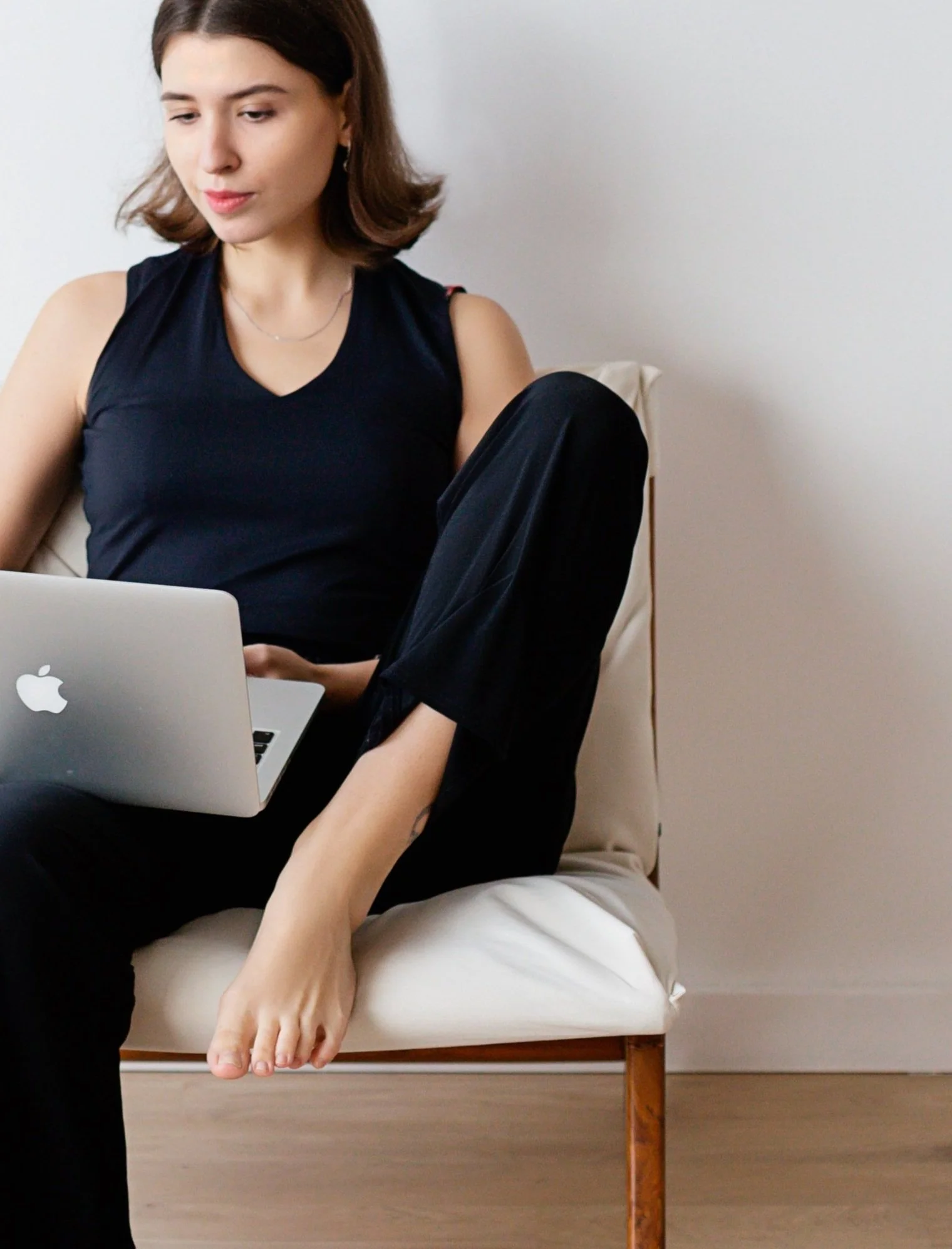 Woman sitting on a white cushioned chair, looking at a laptop on her lap, dressed in black with a neutral expression.