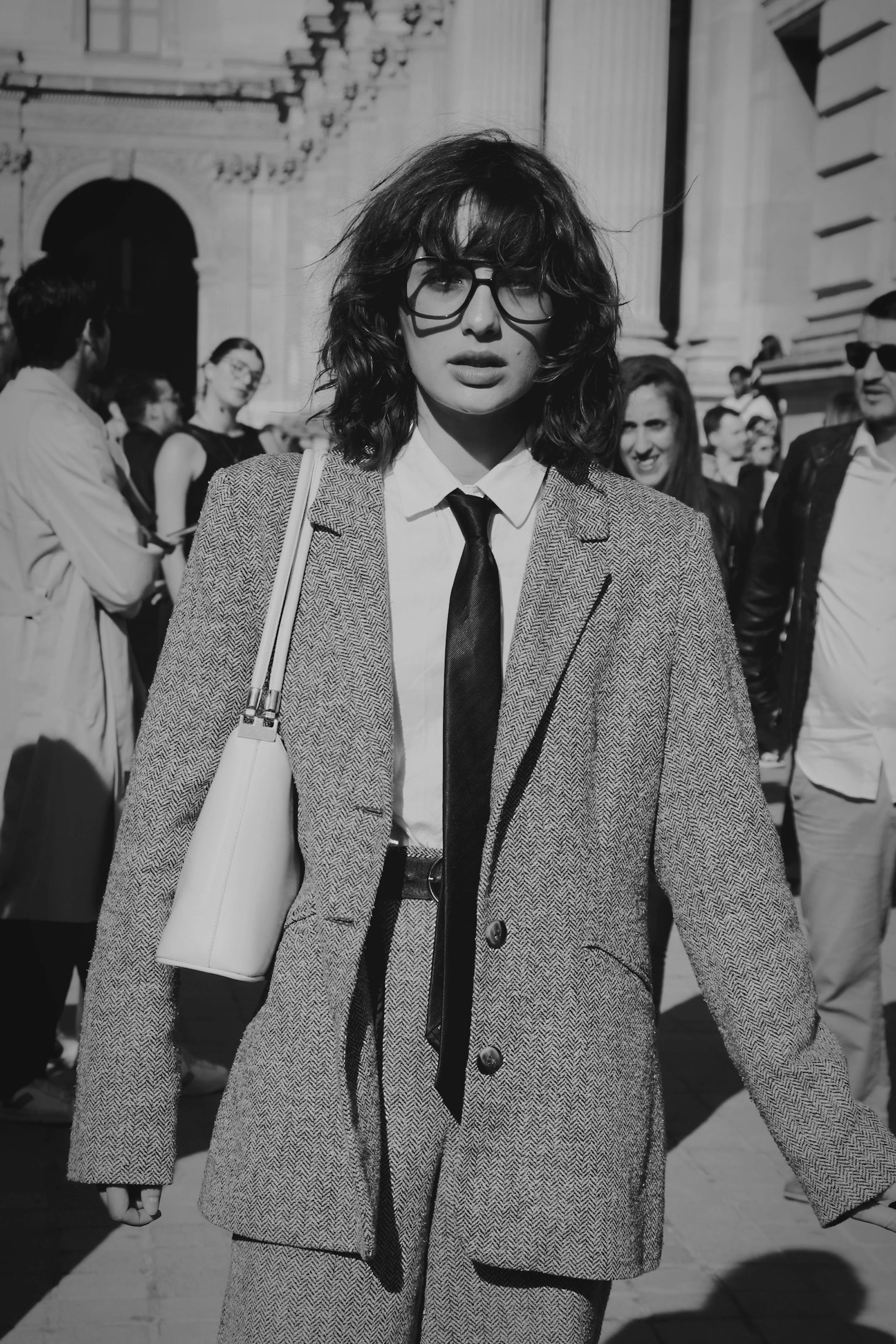 A woman with wavy hair and glasses walking on a city street in sunlight, dressed in a tailored blazer, shirt, and tie, carrying a shoulder bag, with a group of people in the background.