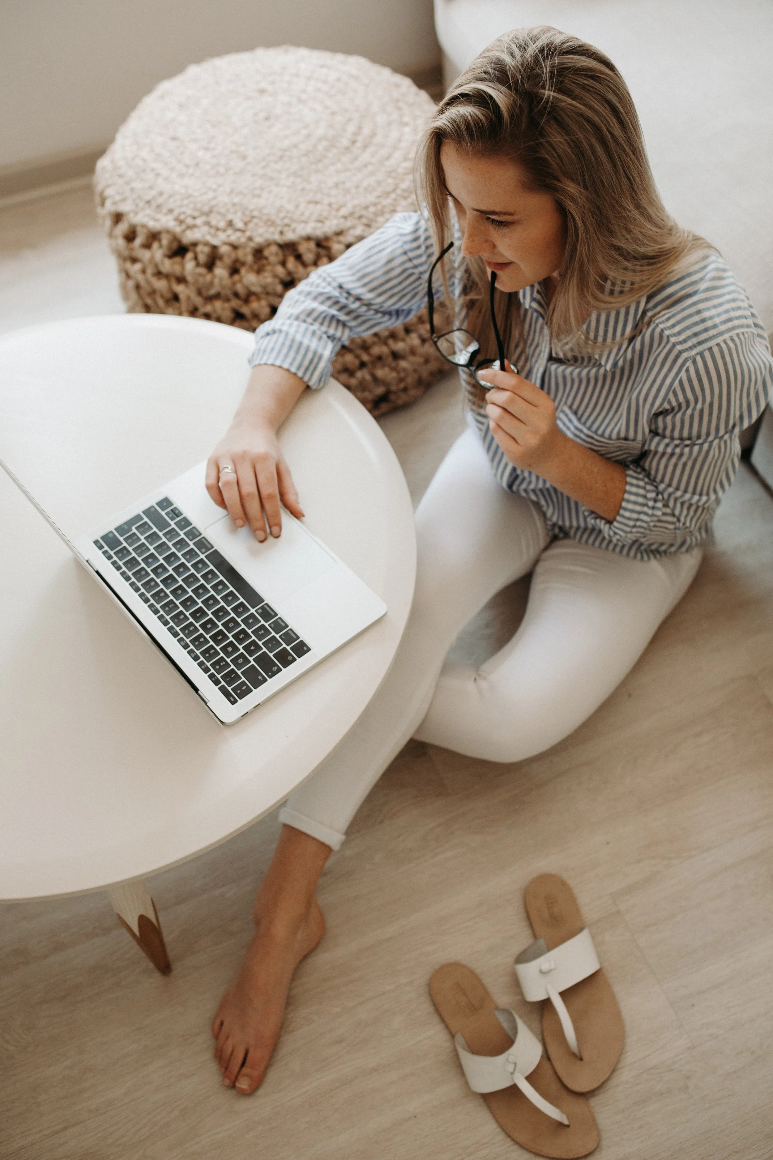 A woman sitting on the floor at a white table, holding glasses and looking at a laptop with a silver body and black keys. She has long, light brown hair and is wearing a blue and white striped shirt and white pants. There is a pair of white sandals on the wooden floor beside her and a textured beige pouf behind her.