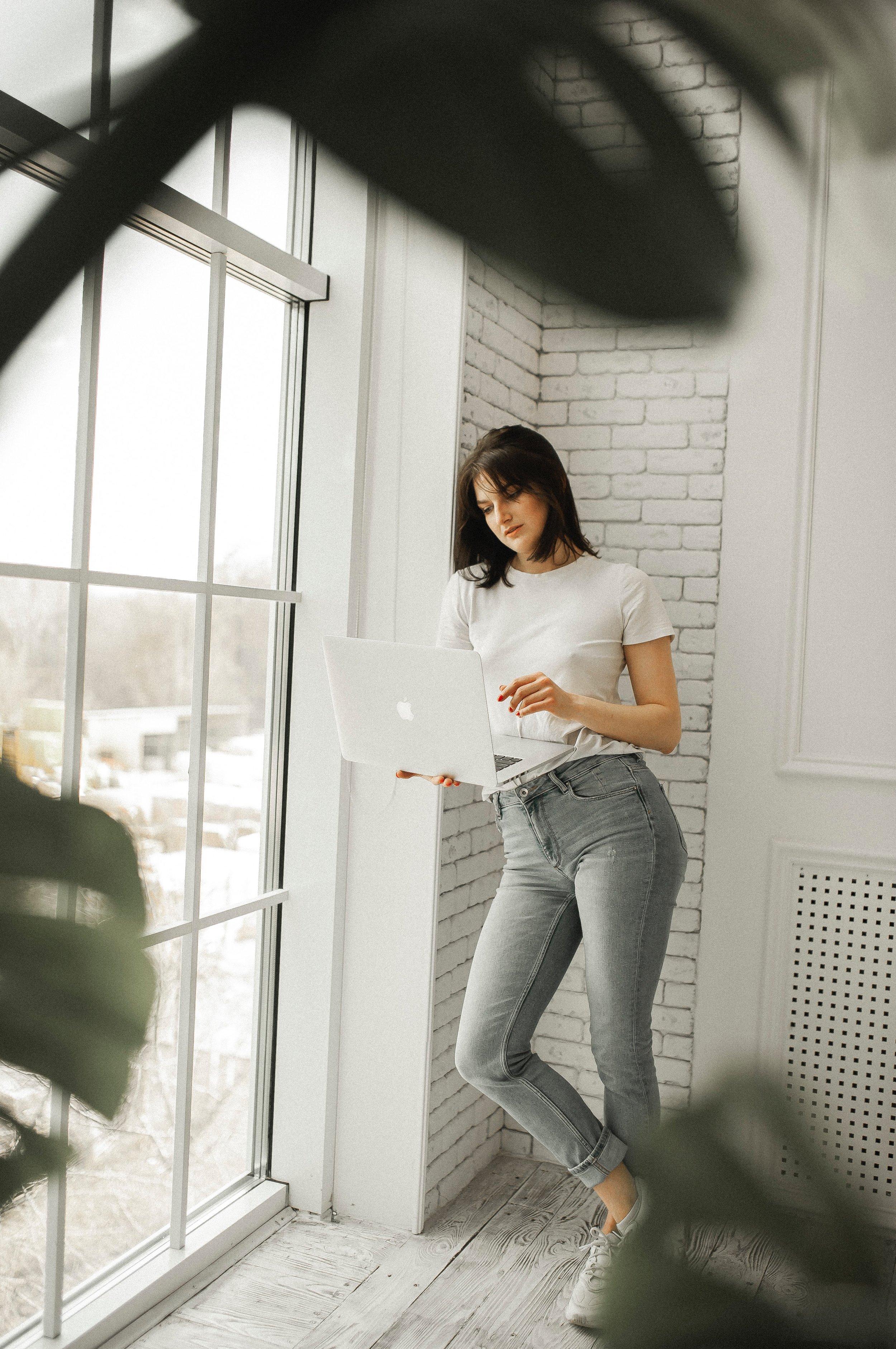 Young woman standing by large window, looking at laptop, with white brick wall behind her