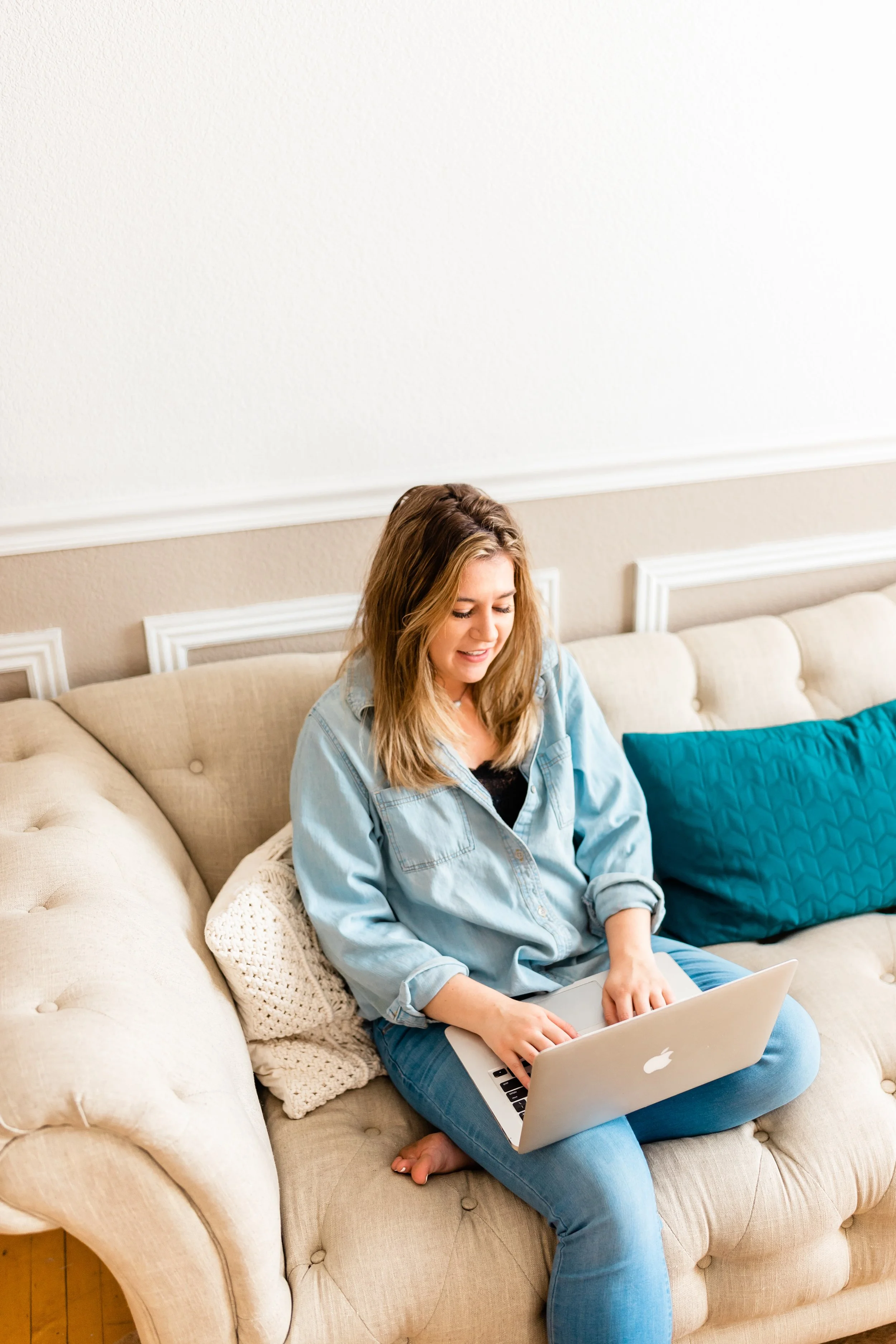 A woman sitting on a beige couch using a silver Apple MacBook laptop.
