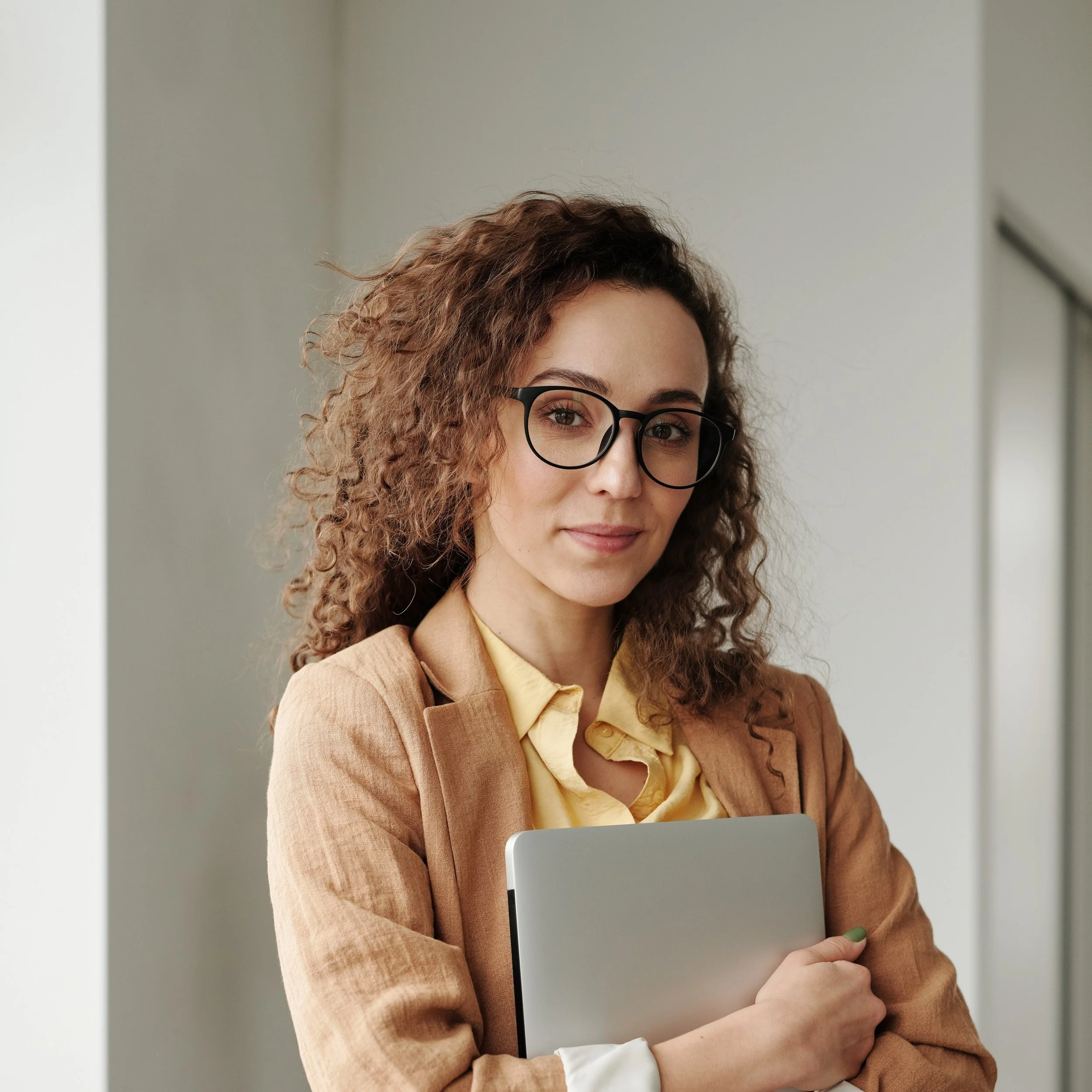 A woman with curly brown hair, glasses, in a tan blazer and yellow shirt, holding a silver laptop, smiling, indoors.